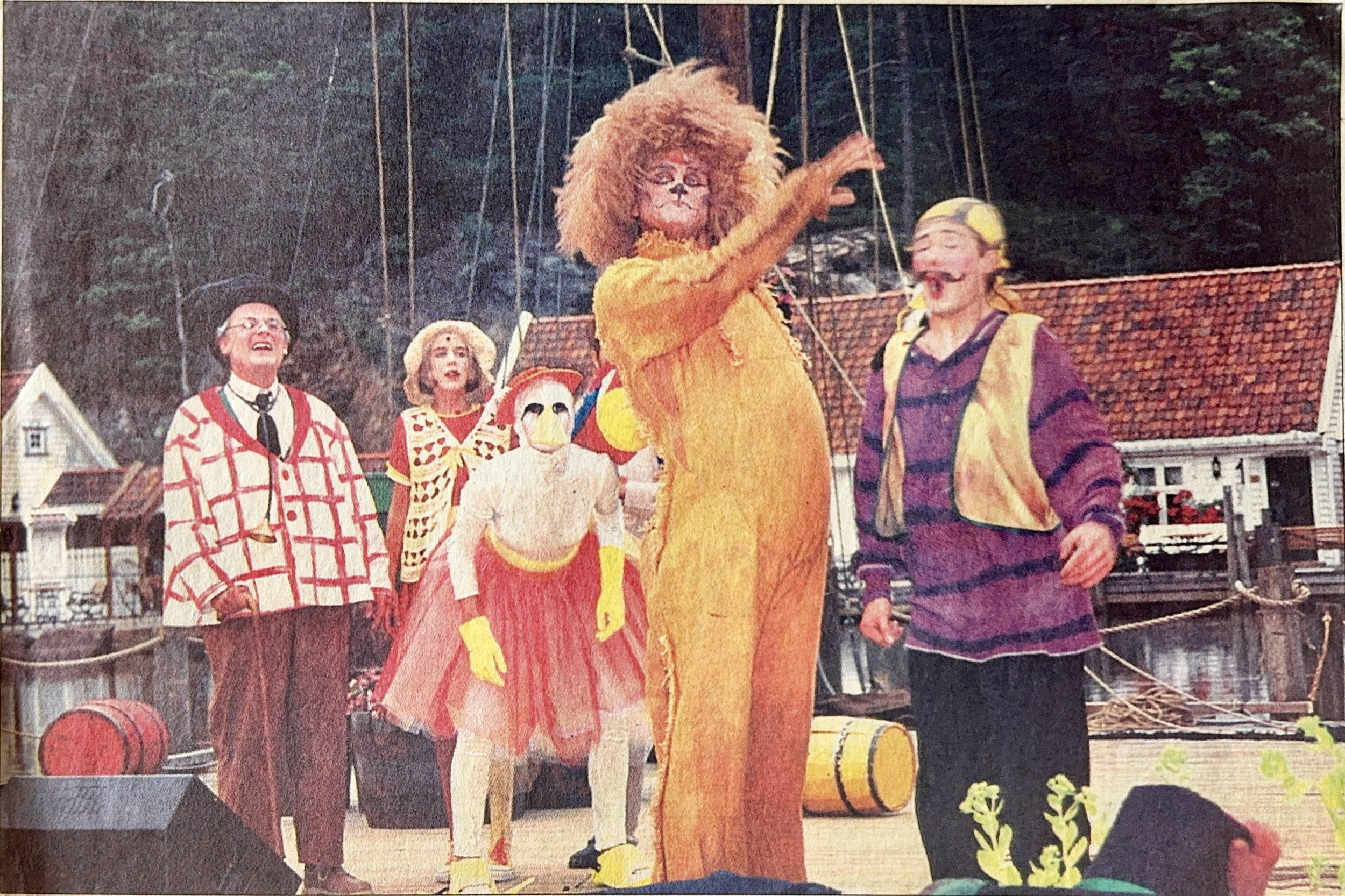 A group of children and adults dressed in colorful costumes, standing outdoors with a backdrop of trees and houses, participating in a festive event or parade.
