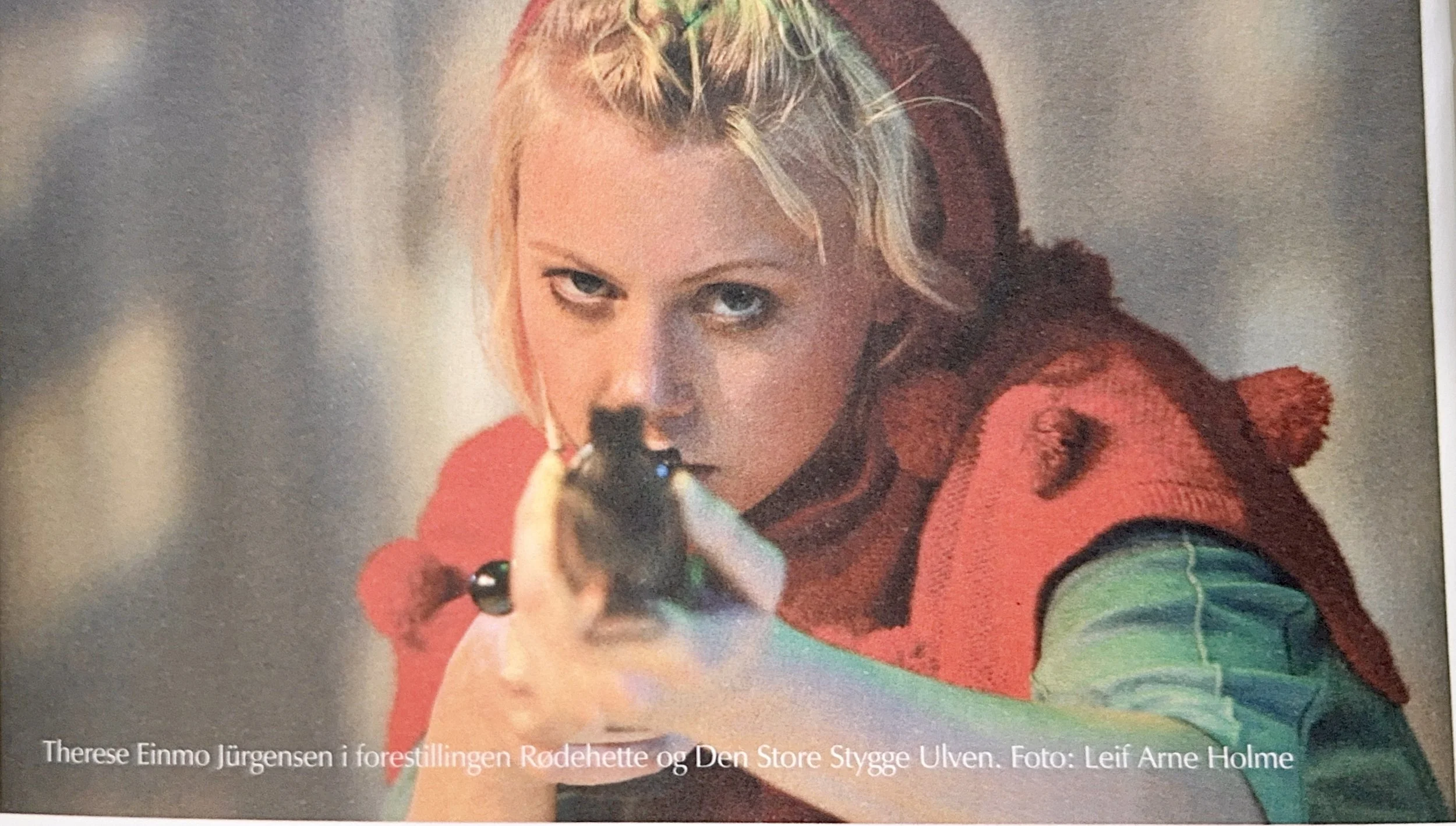A woman in a red and green costume aims a gun directly at the camera, with an intense expression. The background is blurred, bringing focus to her face and outfit.