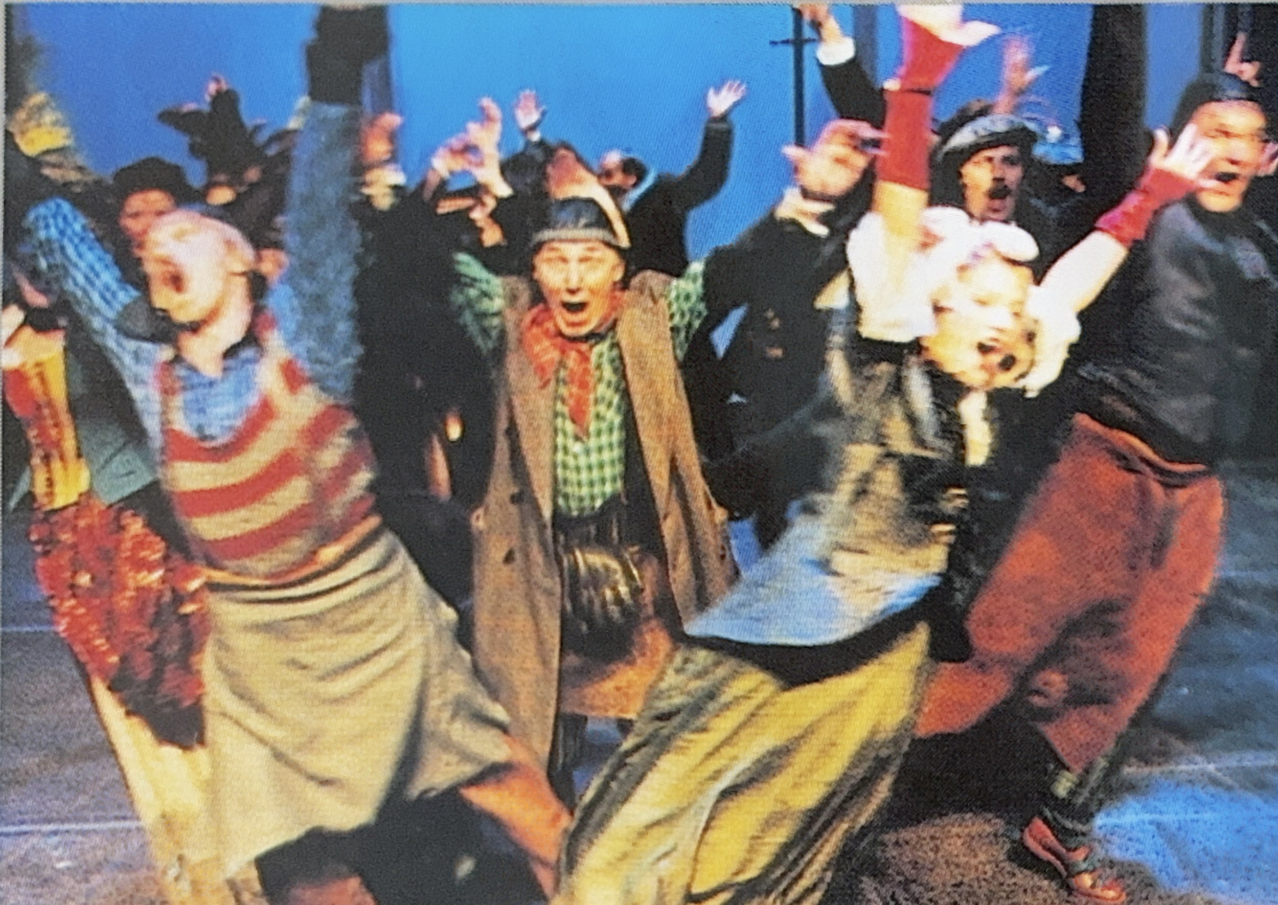 A group of people dressed in festive costumes, celebrating and smiling, with their hands raised or making joyful gestures, against a blue background.