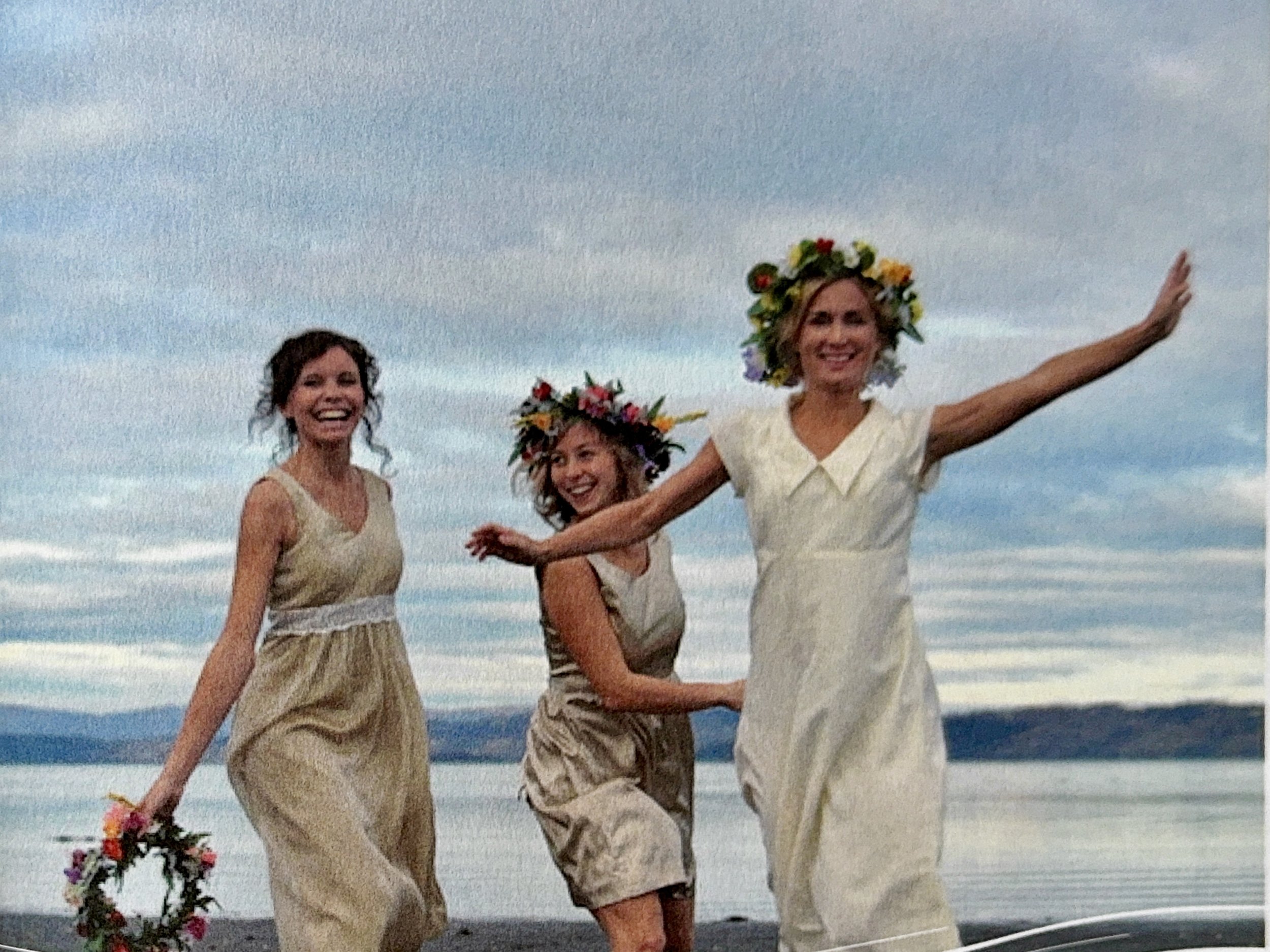 Three women with floral crowns and beige dresses standing by the water at sunset, smiling and celebrating.