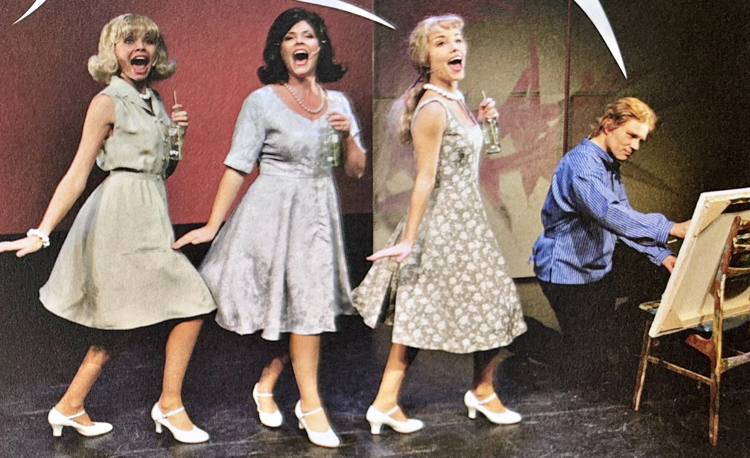 Four women in vintage dresses and white heels singing and dancing together, with one man playing the piano in the background.