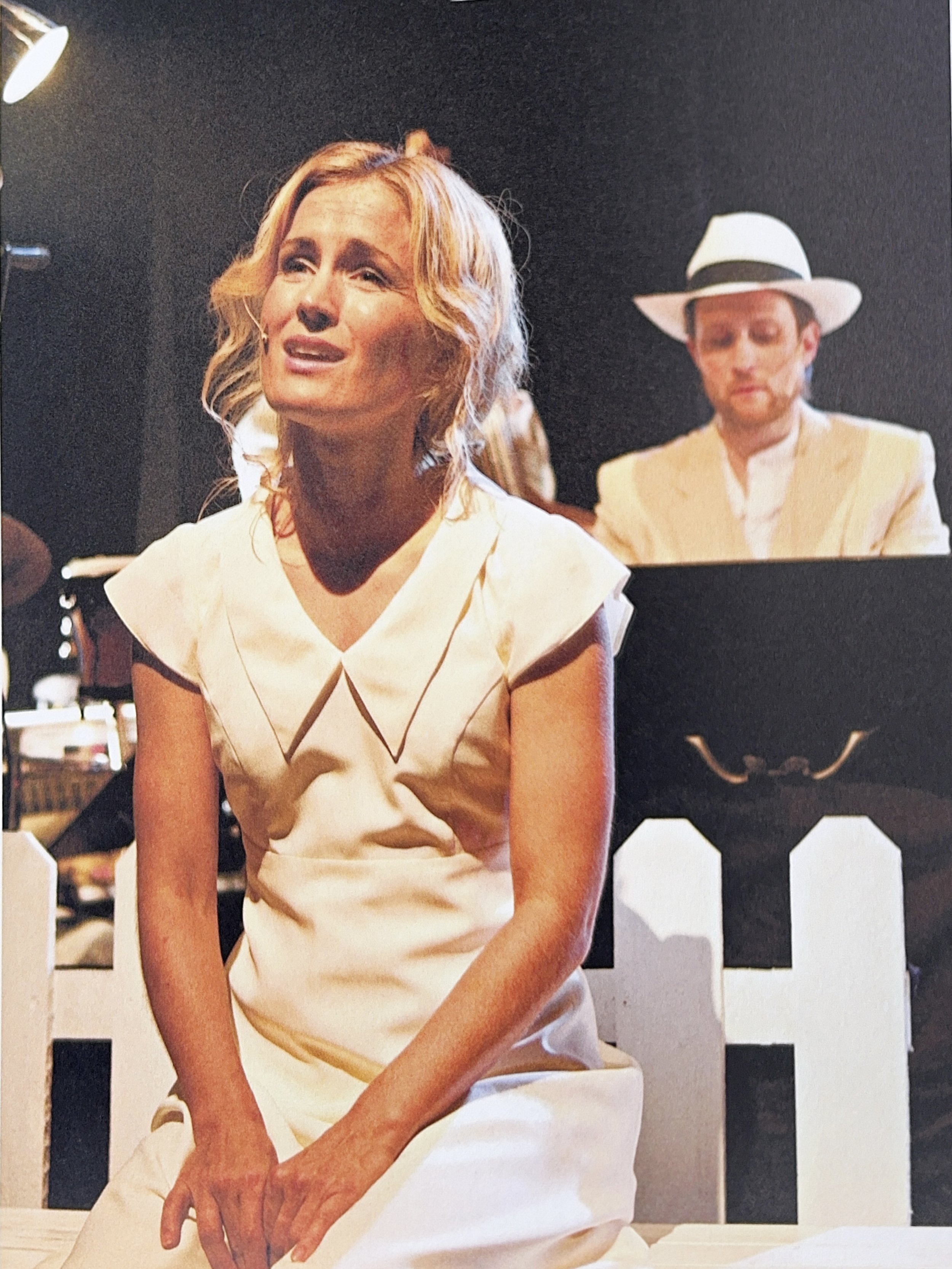 A woman with blonde wavy hair, wearing a white dress, sitting at a table with her hands folded. Behind her, a man in a white hat and a yellow outfit is visible, slightly out of focus. The background is dark and the setting appears to be indoors.