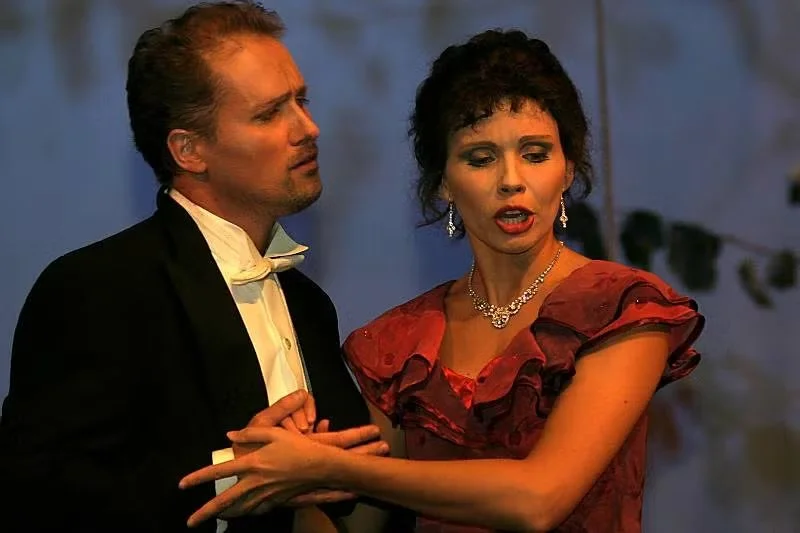 Two actors on stage, a man in formal tuxedo and a woman in a red dress, seemingly in an emotional or dramatic scene.