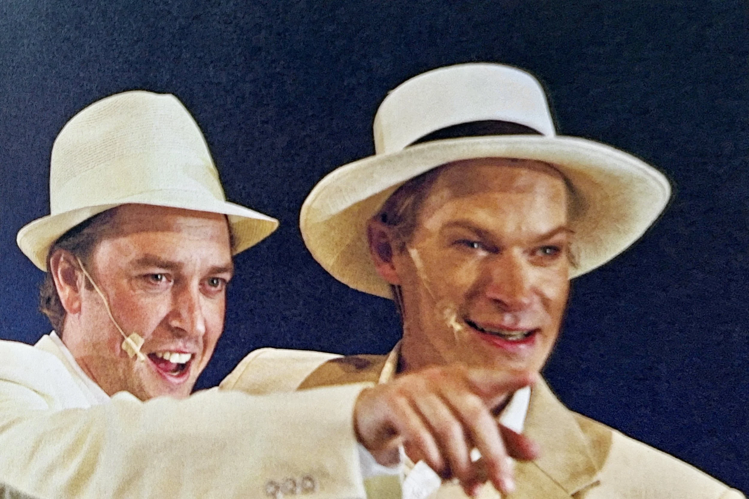 Two men wearing white suits and wide-brimmed white hats smiling and posing for a photo against a dark background.