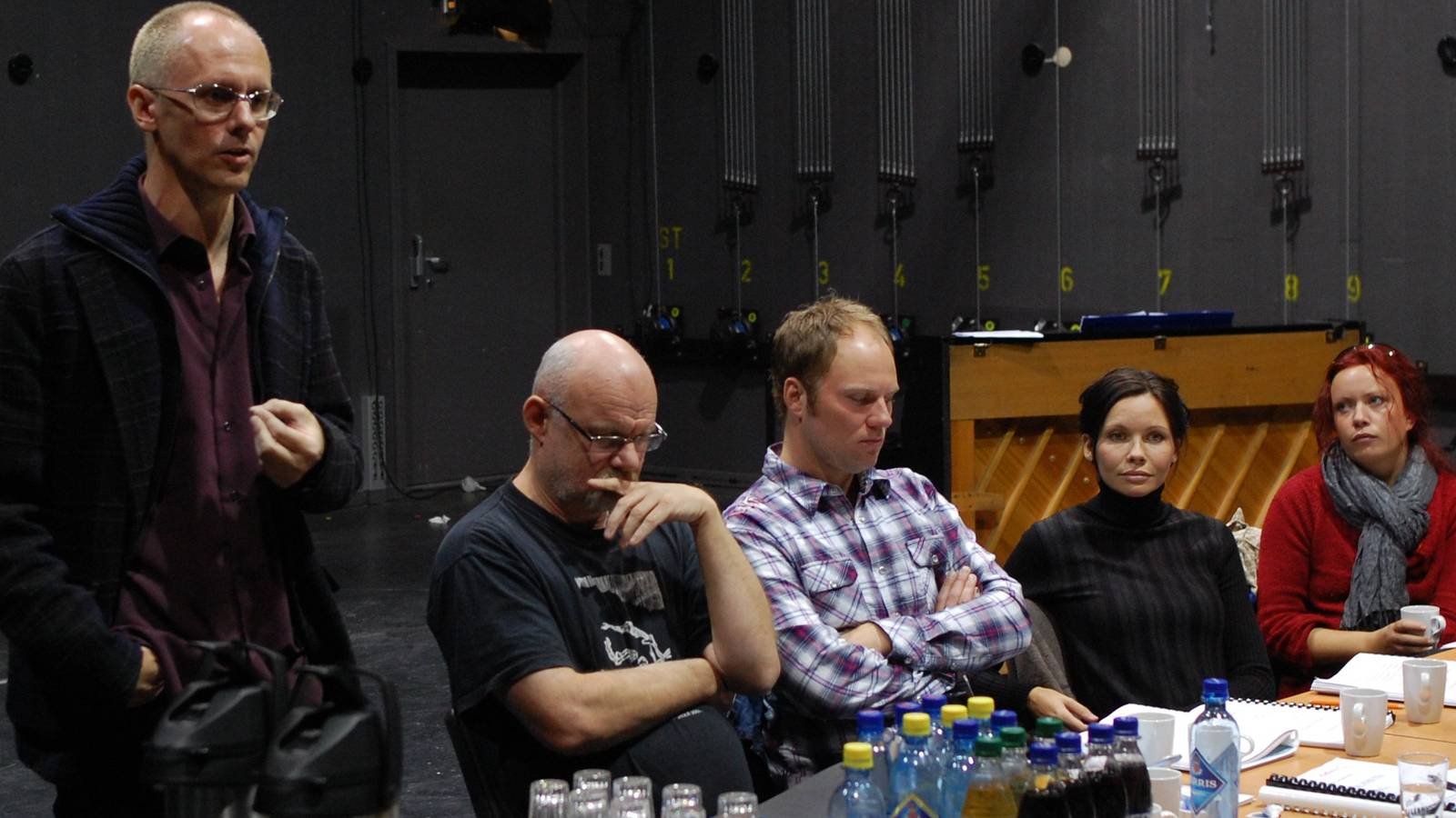 A group of five people sitting at a table with drinks and notebooks, and one man standing, in a dark room with equipment on the wall.