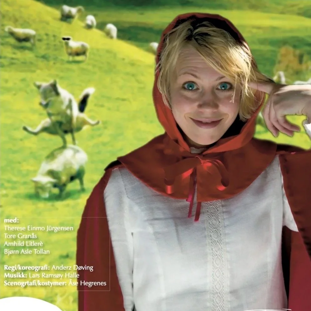 A woman dressed as Little Red Riding Hood with a red cape and hood, smiling and posing with her hand to her ear, in front of a green landscape with sheep.