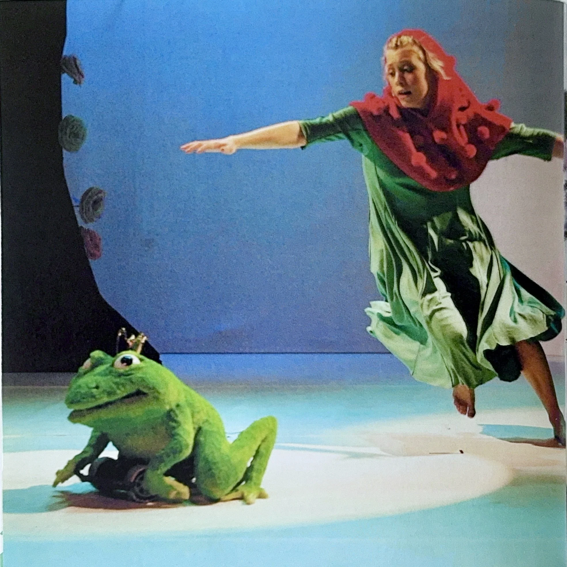 A woman with a red hoodie and green clothes doing a split on the ground, while a green plush frog watches.
