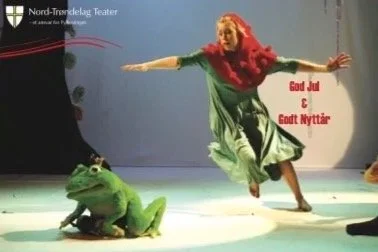 A person with short blonde hair in a red shirt and green skirt performing on a stage with a large frog costume. There is text in Swedish wishing a Merry Christmas and Happy New Year.