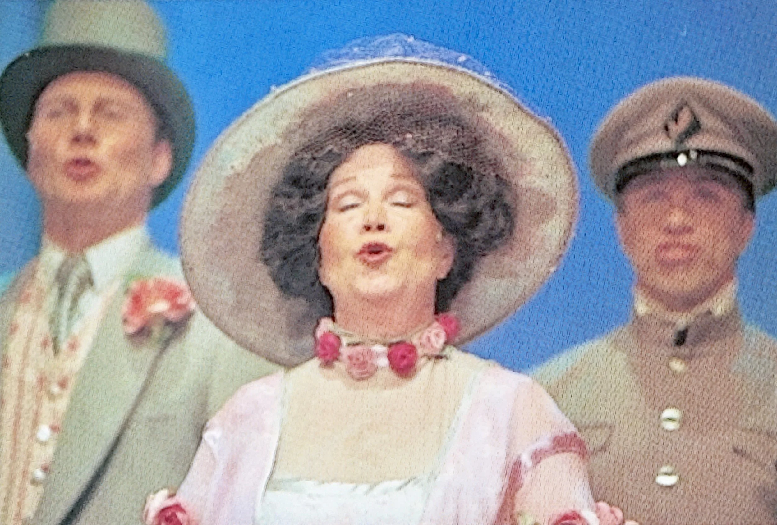 A vintage color photograph of three children in old-fashioned clothing and hats, smiling and making faces against a bright blue background.