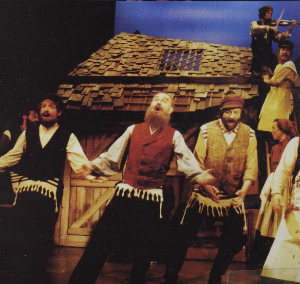 Actors performing in a theatrical play with a rustic wooden house backdrop and a stage. Some performers are singing or dancing, dressed in period costumes.