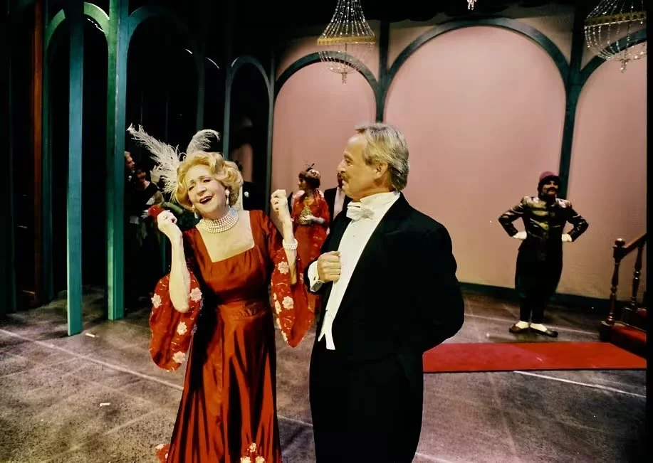 Two people in elegant attire dancing and laughing at a formal event in a decorated ballroom with chandeliers.