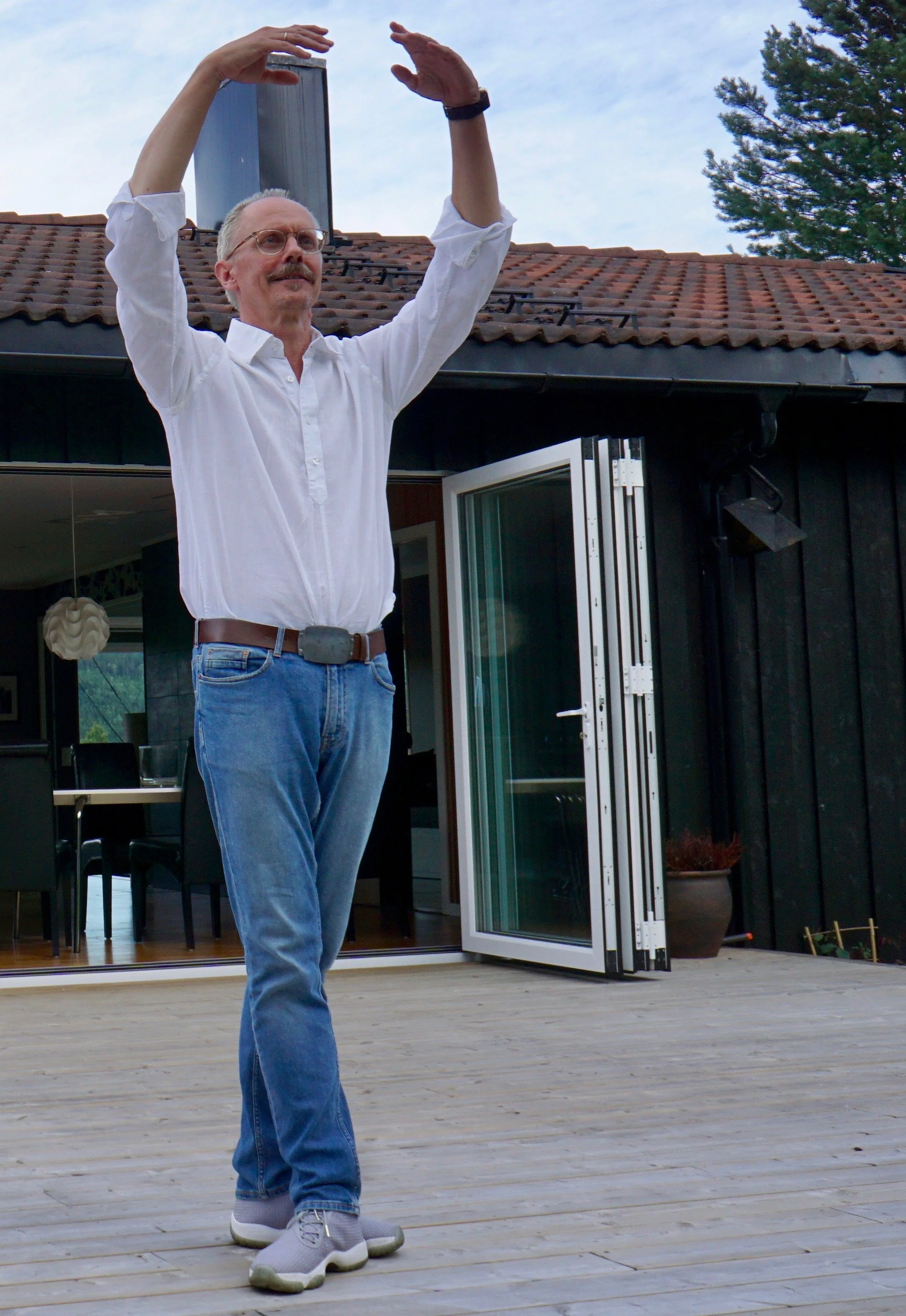 A man with glasses, a white shirt, and jeans standing on a wooden patio, stretching his arms above his head outdoors, with a house and trees in the background.