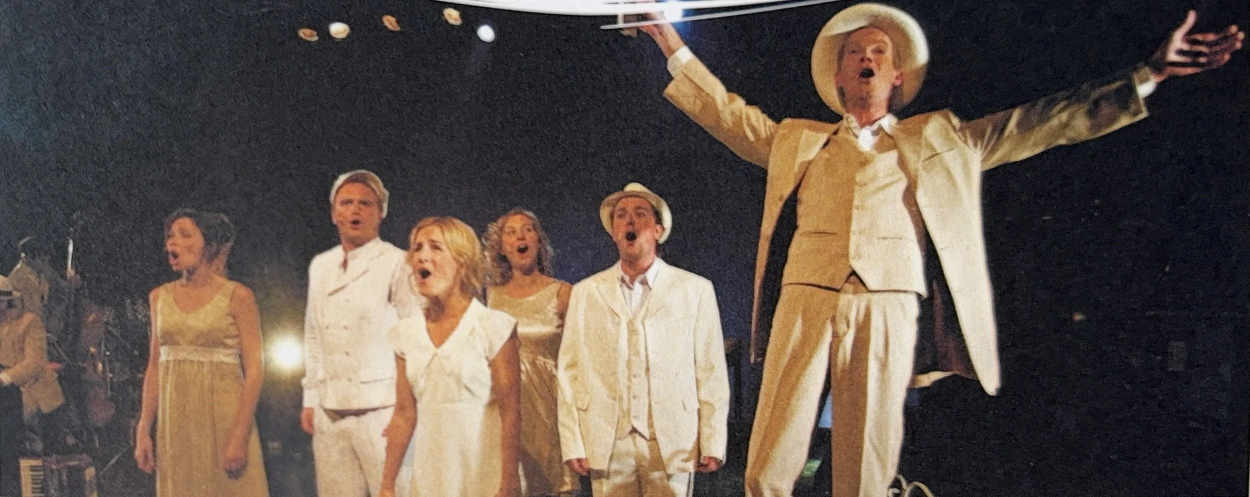 A group of people singing on stage at night, with one person in front with outstretched arms, all dressed in white and beige outfits.