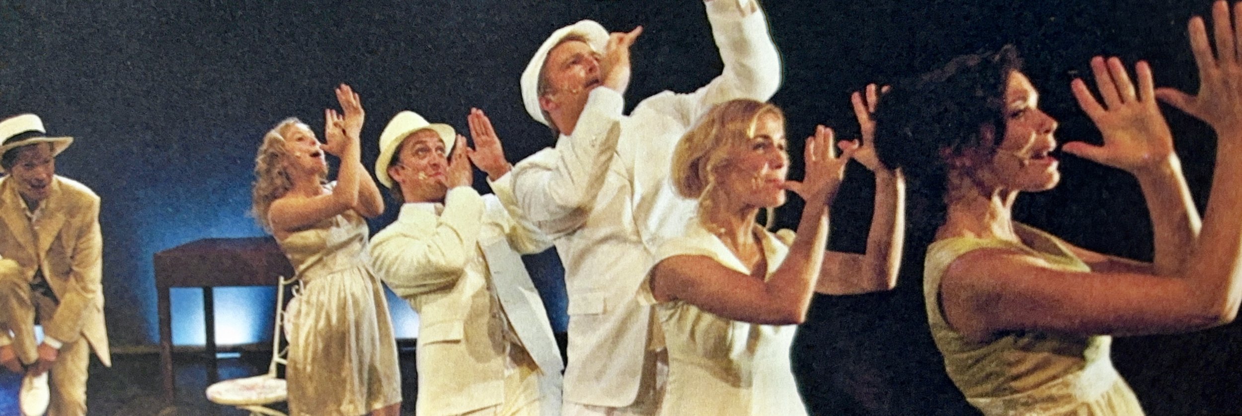 A group of actors on stage dressed in vintage white and light-colored clothing, with some wearing hats, performing in a theatrical production with animated gestures and facial expressions.