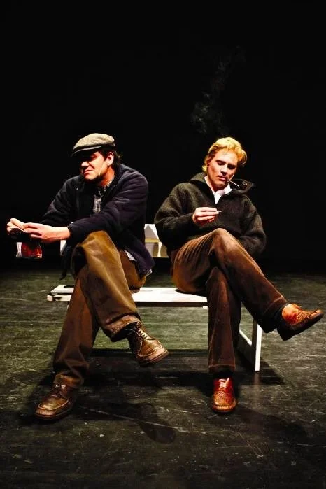 Two men sitting on a bench, both looking at their phones, with a dark background, one wearing a cap and the other with blond hair, smoking.
