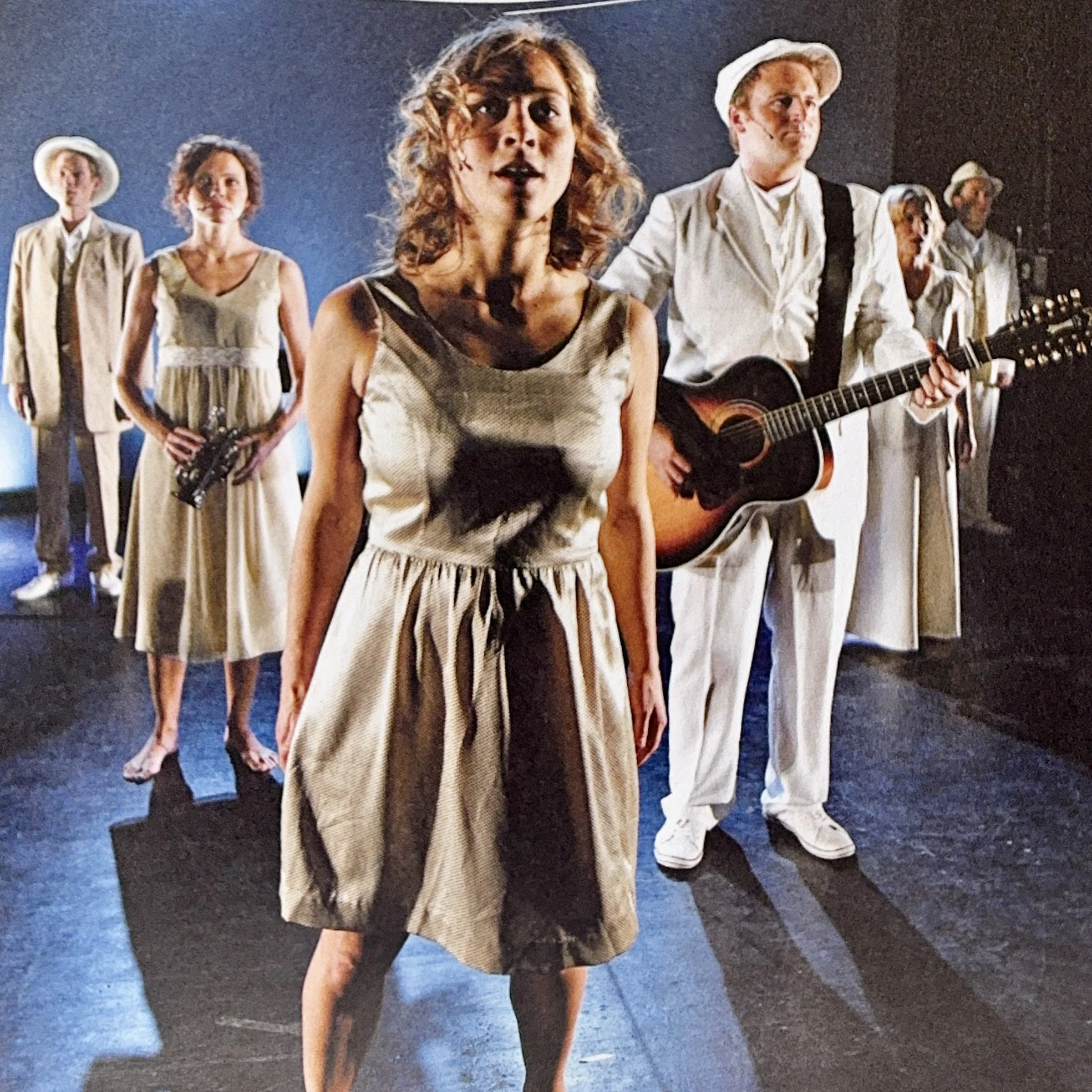 A group of people standing in a dark room, with one woman in the foreground holding a microphone and another woman in the background holding a saxophone. The woman in the foreground is wearing a sleeveless top and the woman holding the saxophone is d