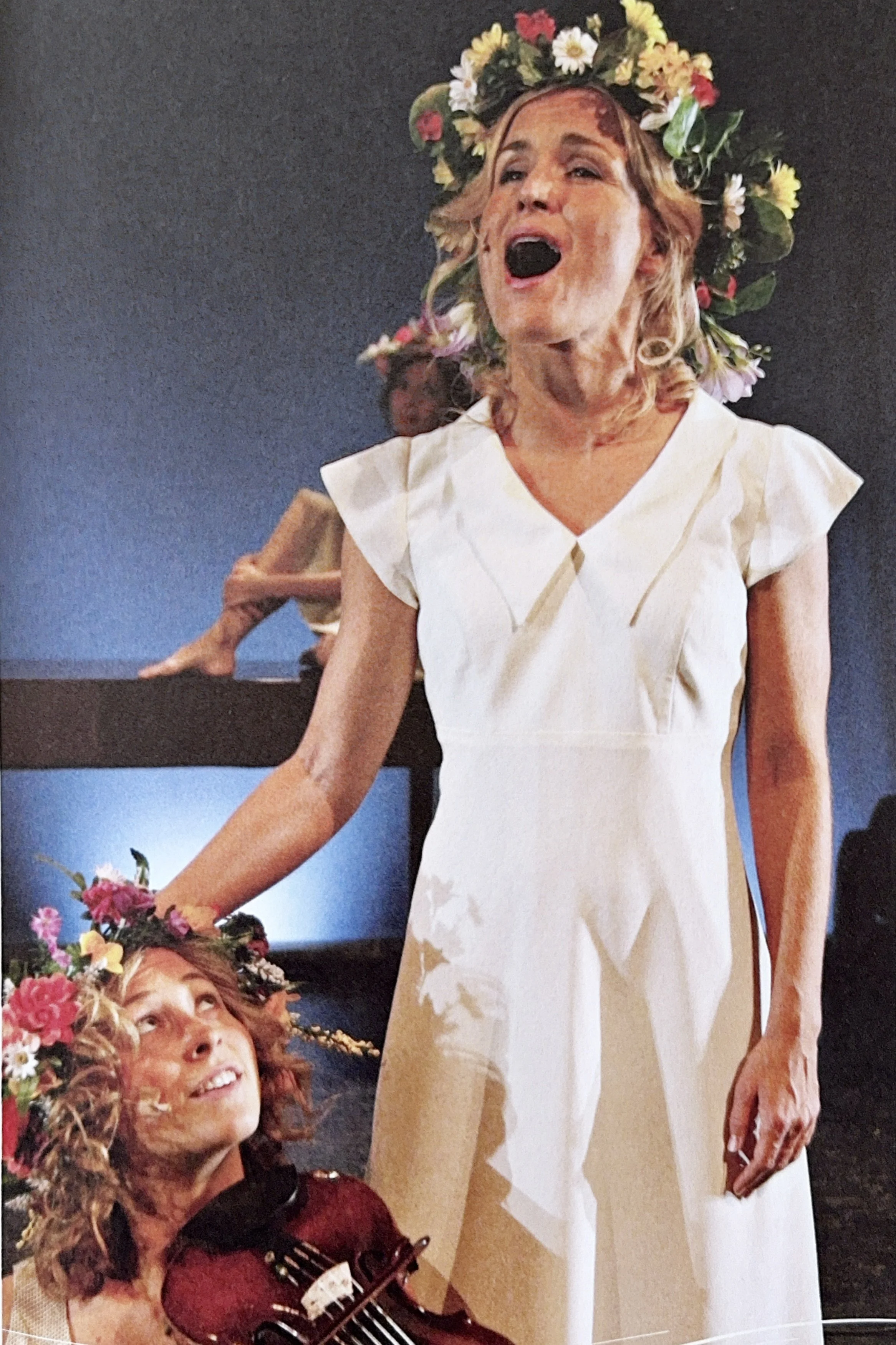 Two women, one with curly hair wearing a floral crown and a white dress, singing and surprised, and the other with curly hair wearing a floral crown and playing ukulele, looking up and smiling.