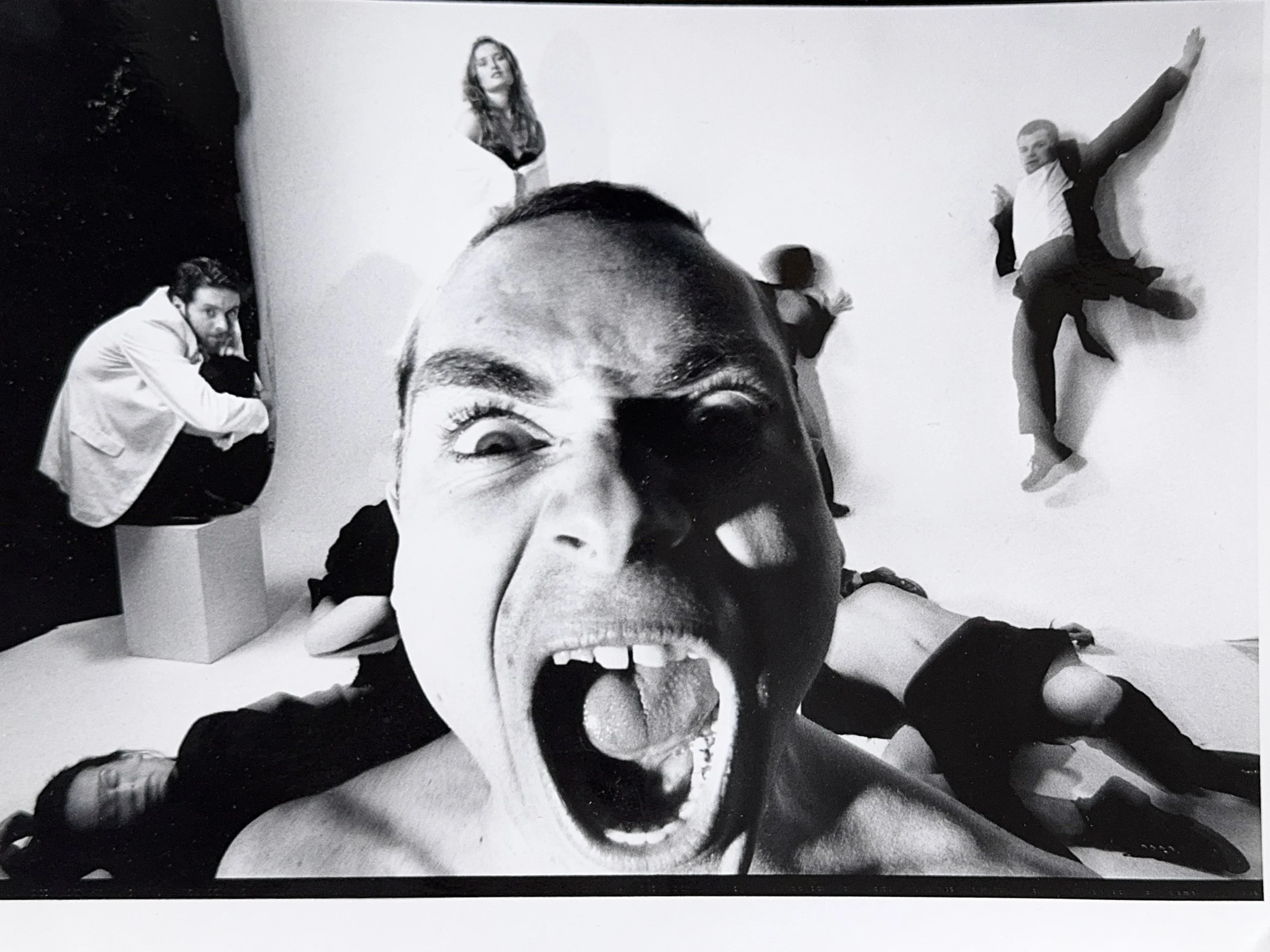Black-and-white photograph of a wall with a large face portrait of a woman with her tongue out. Several people are interacting with the artwork, with some climbing or leaning against the wall, including a man on the right in a suit and tie, a woman in the middle, and another man on the left side.