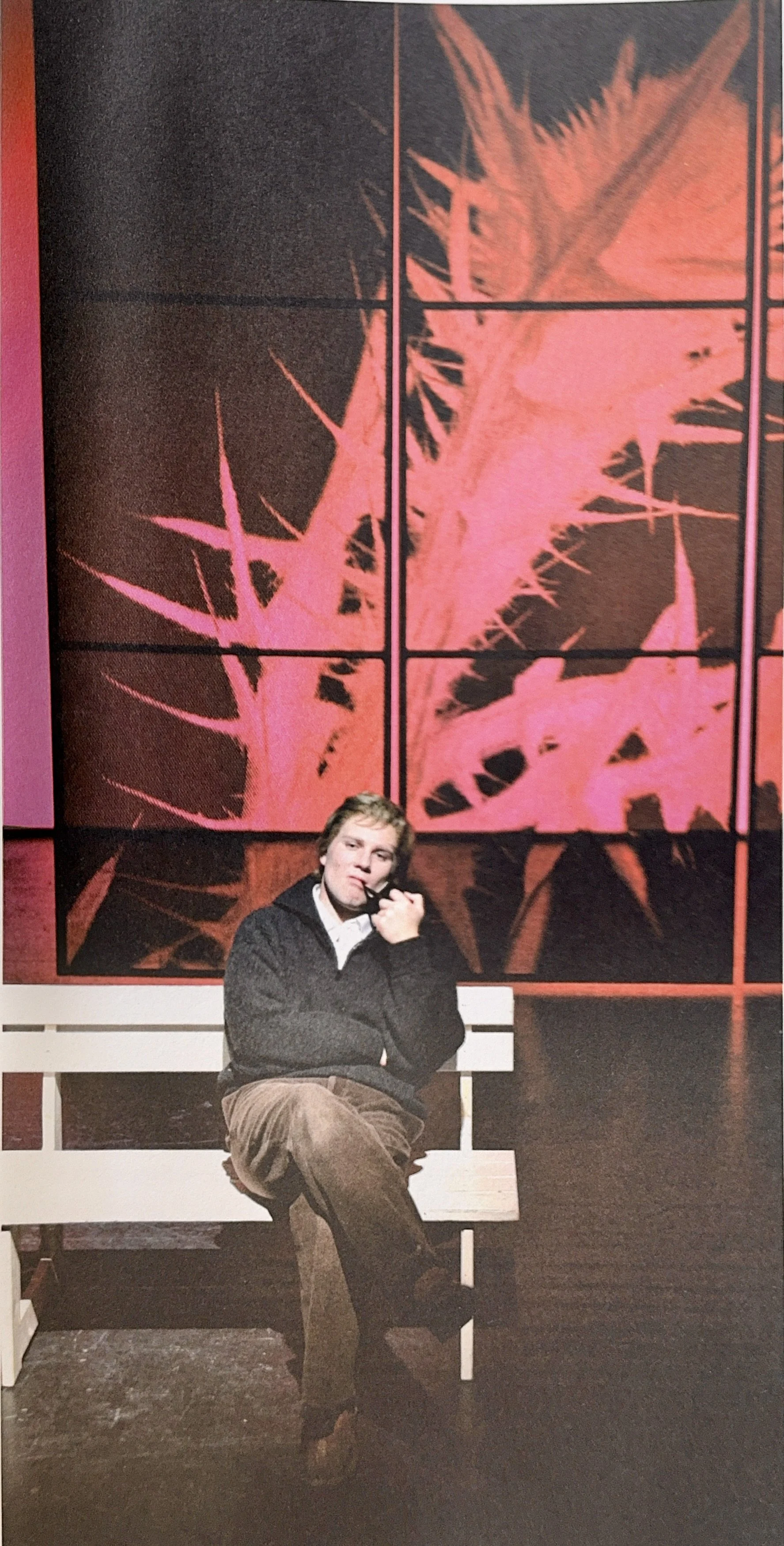 A young man with light brown, slightly curly hair sitting on a white bench, wearing a black sweater over a white shirt and brown pants. He is holding a pipe to his mouth with his right hand, appearing to be in thought, with a large abstract artwork w