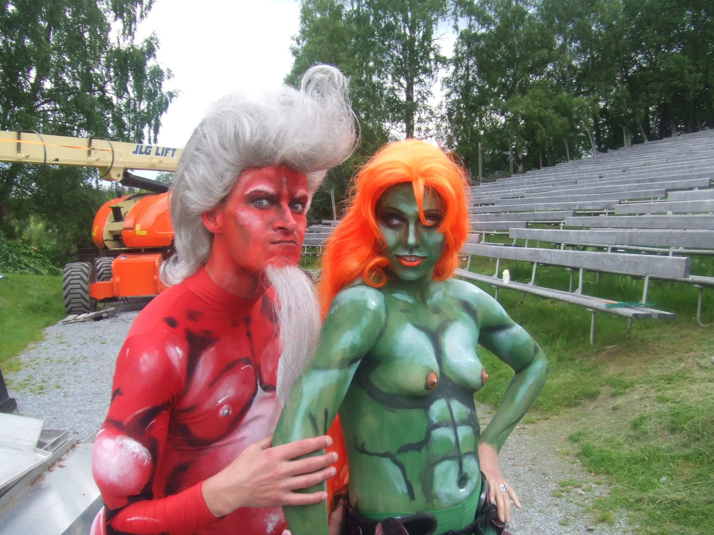 Two people dressed as comic book characters, one as a red and white themed character with a white wig and beard, and the other as a green and orange character with green face paint and bright orange hair, standing outdoors near metal bleachers and a construction lift.