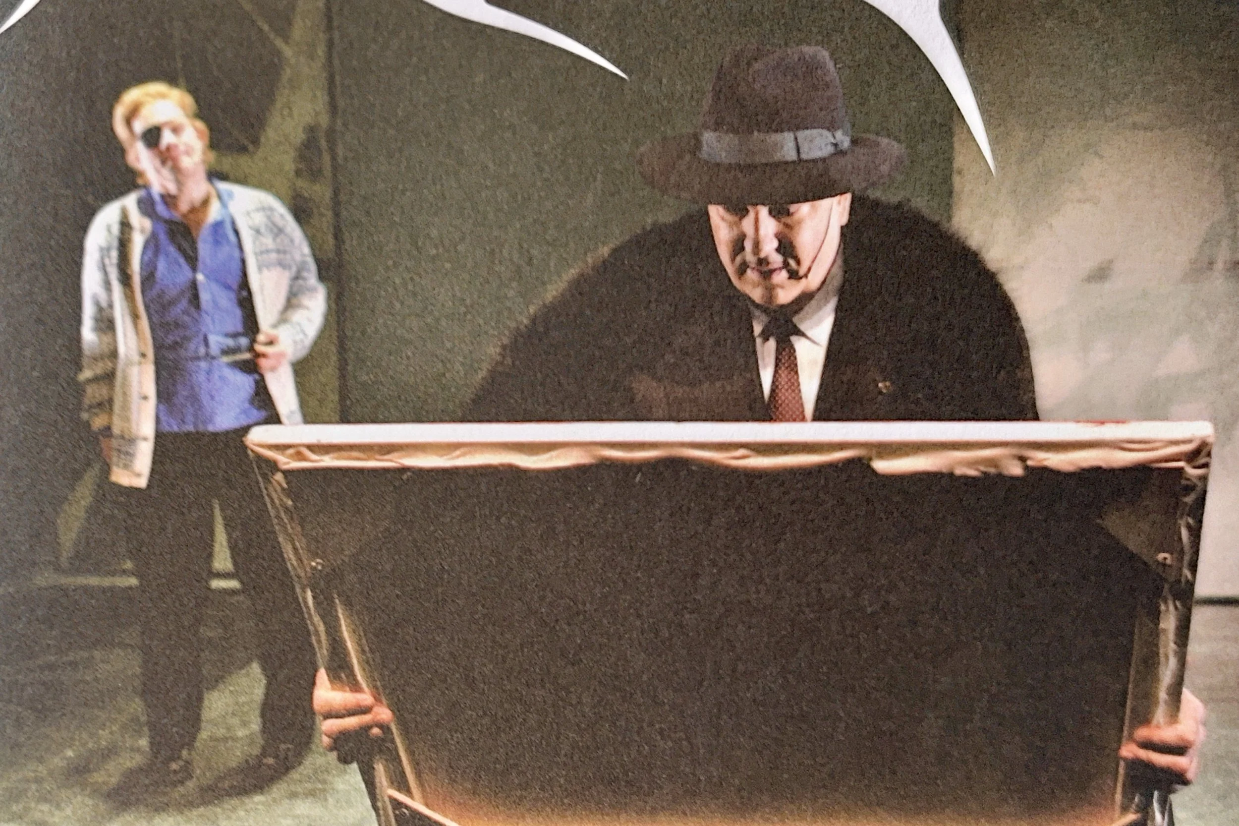 Two men, one in a blue shirt and beige jacket, and the other in a black suit with a brown hat and red tie, are standing near a basket with a large white umbrella overhead in an outdoor setting at night.