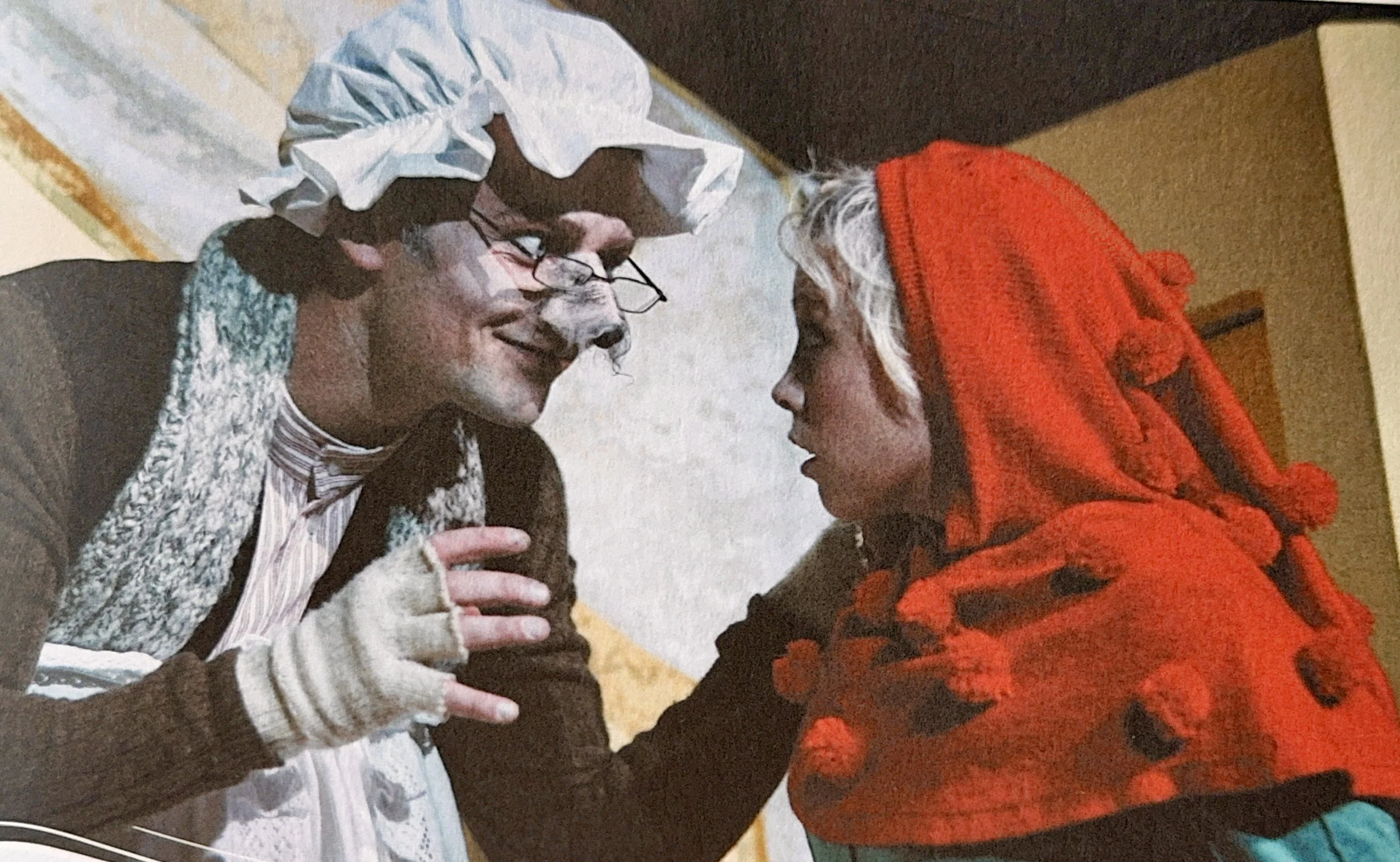 Two people are sitting close together and smiling at each other; one is wearing glasses, a striped shirt, and a cozy sweater, while the other is wearing a red hoodie. They appear to be enjoying a moment of conversation or connection.