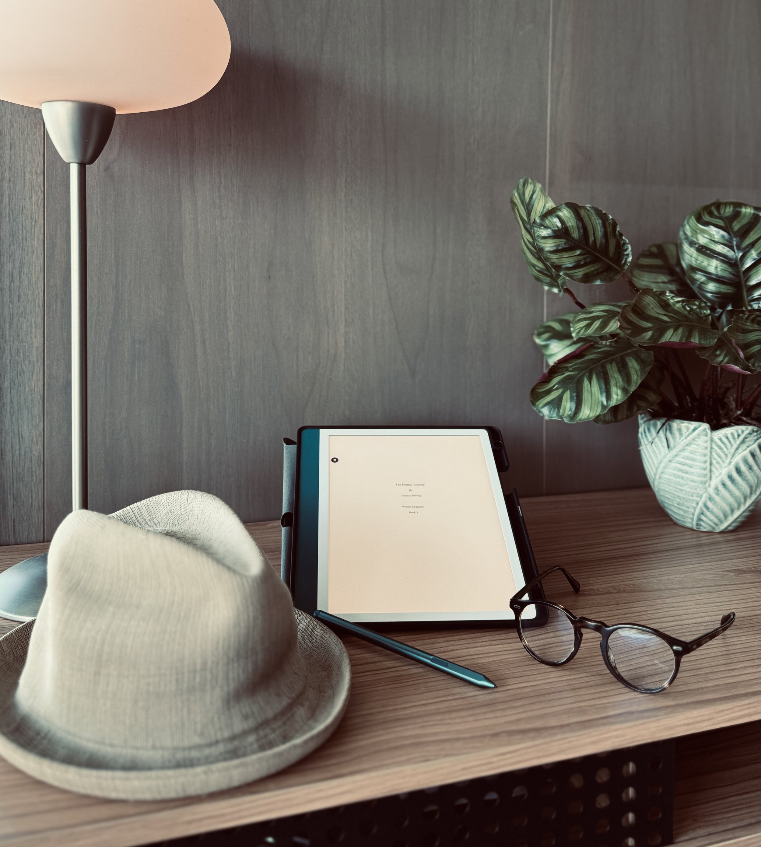 A beige wide-brimmed hat, black eyeglasses, and a blue pen are placed on a wooden table. The table also holds an electronic device displaying a document titled "The Essential Journey" with the subtitle "Anxiety Claring" and "Women's Endurance." In the background, there's a modern floor lamp with a beige shade and a green plant with large variegated leaves.
