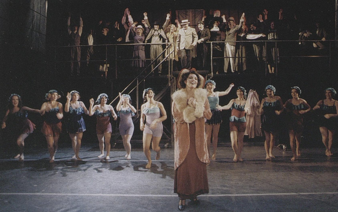 A theatrical stage scene from a musical, featuring a woman in vintage attire in front of a chorus of dancers dressed in colorful costumes with some in lingerie, with an audience in the background.