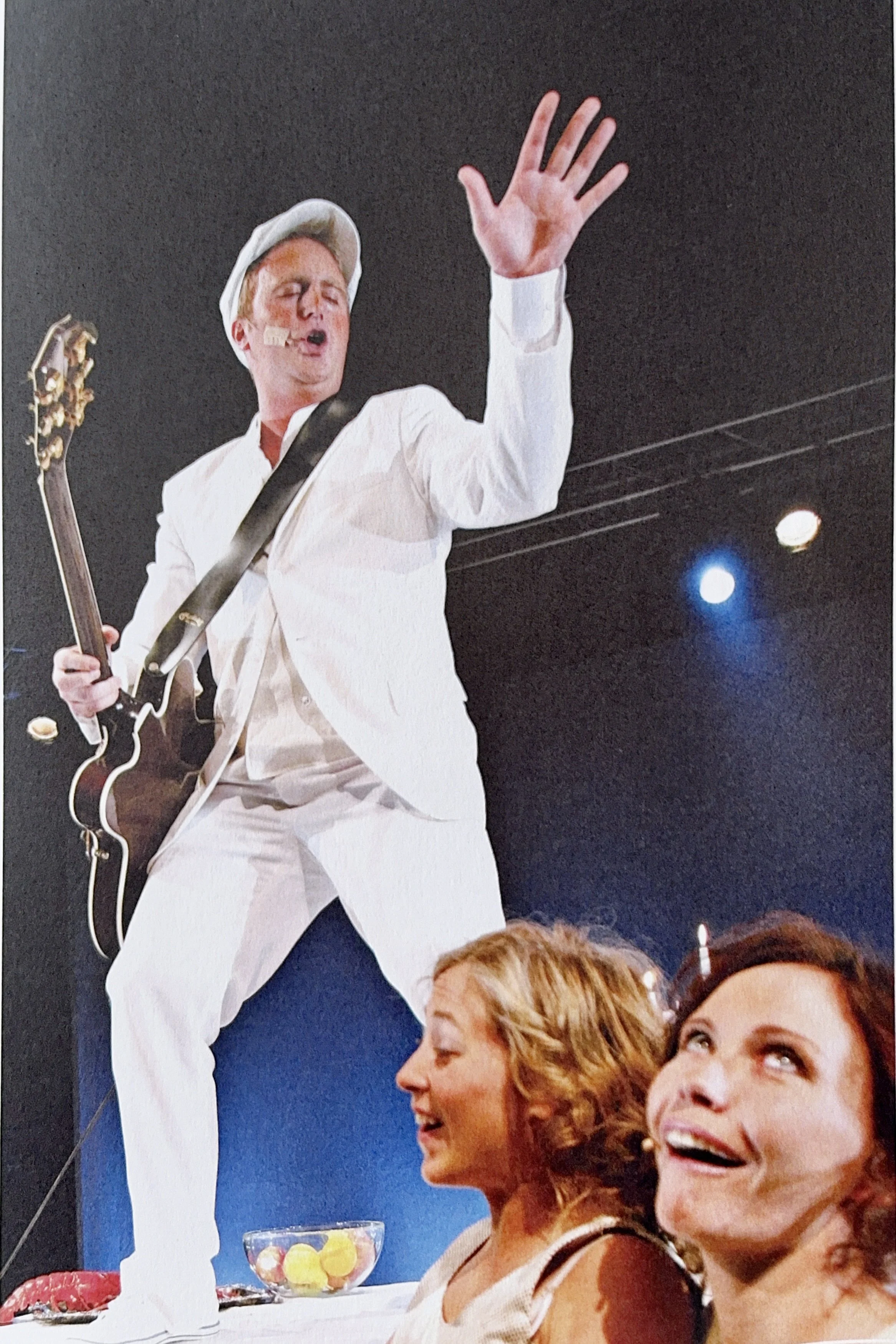 A man playing guitar and singing into a microphone on stage, with a white hat and white outfit, next to a black background with stage lights. Two women are seated, smiling and looking up at him.