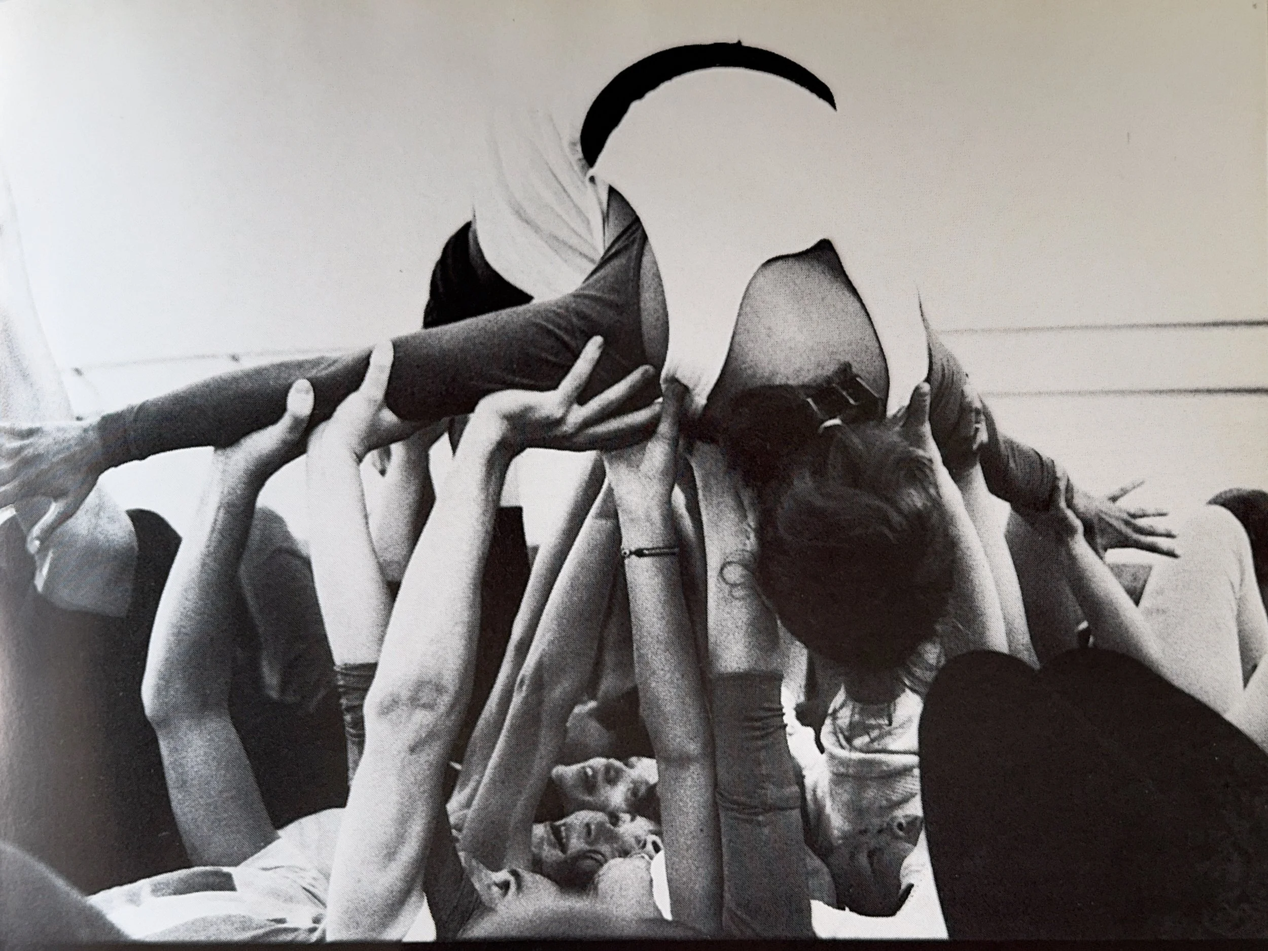 Black and white photo showing multiple hands reaching towards and touching a person lying on the ground, with some hands holding a hat.