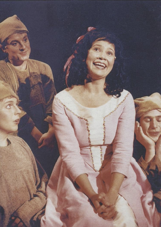 A woman with curly black hair and a red ribbon is smiling, sitting in front of three men dressed in medieval costumes, on a black background.