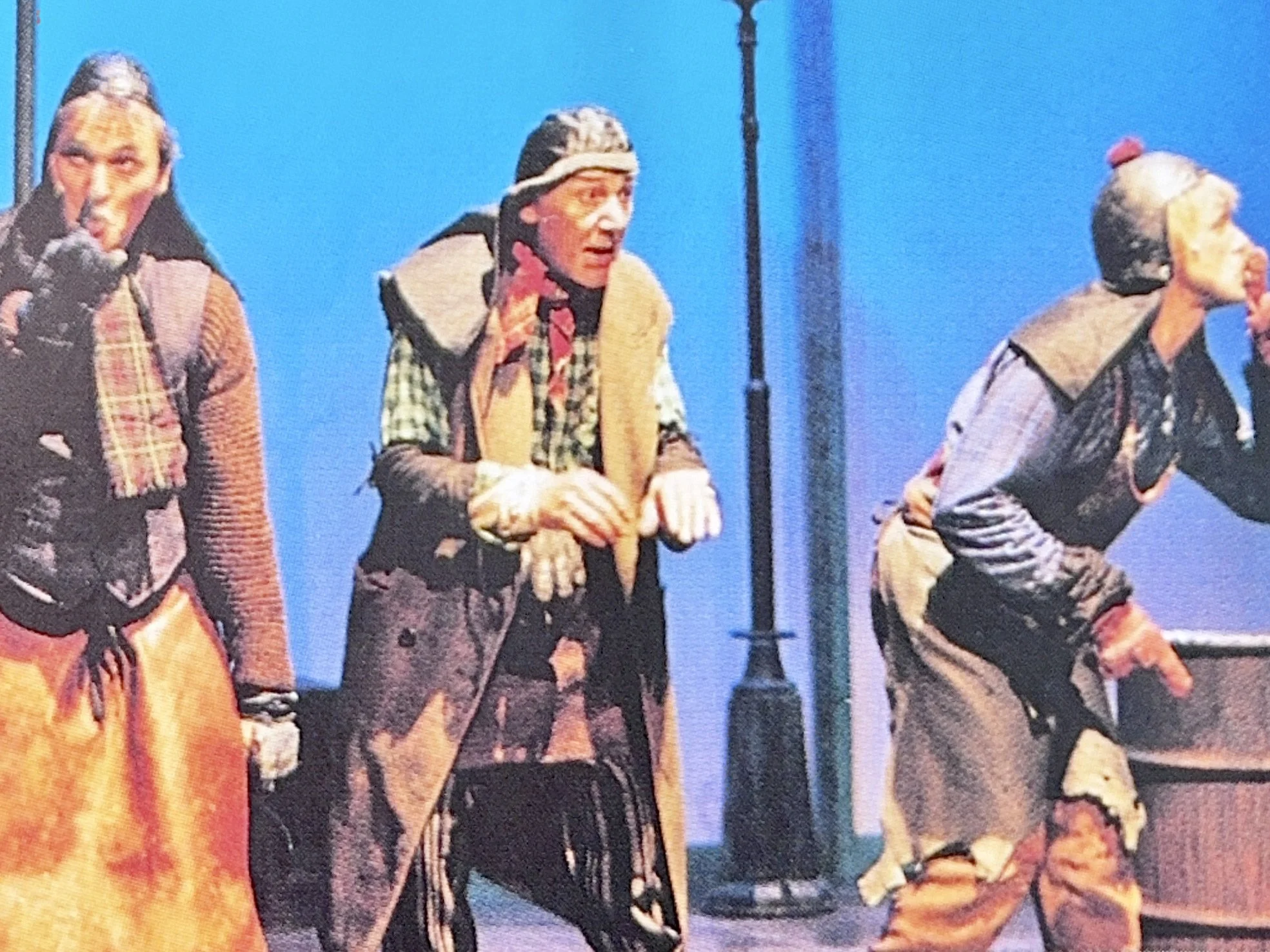 Three performers on stage in costume, with a blue background, engaging the audience with expressive gestures.