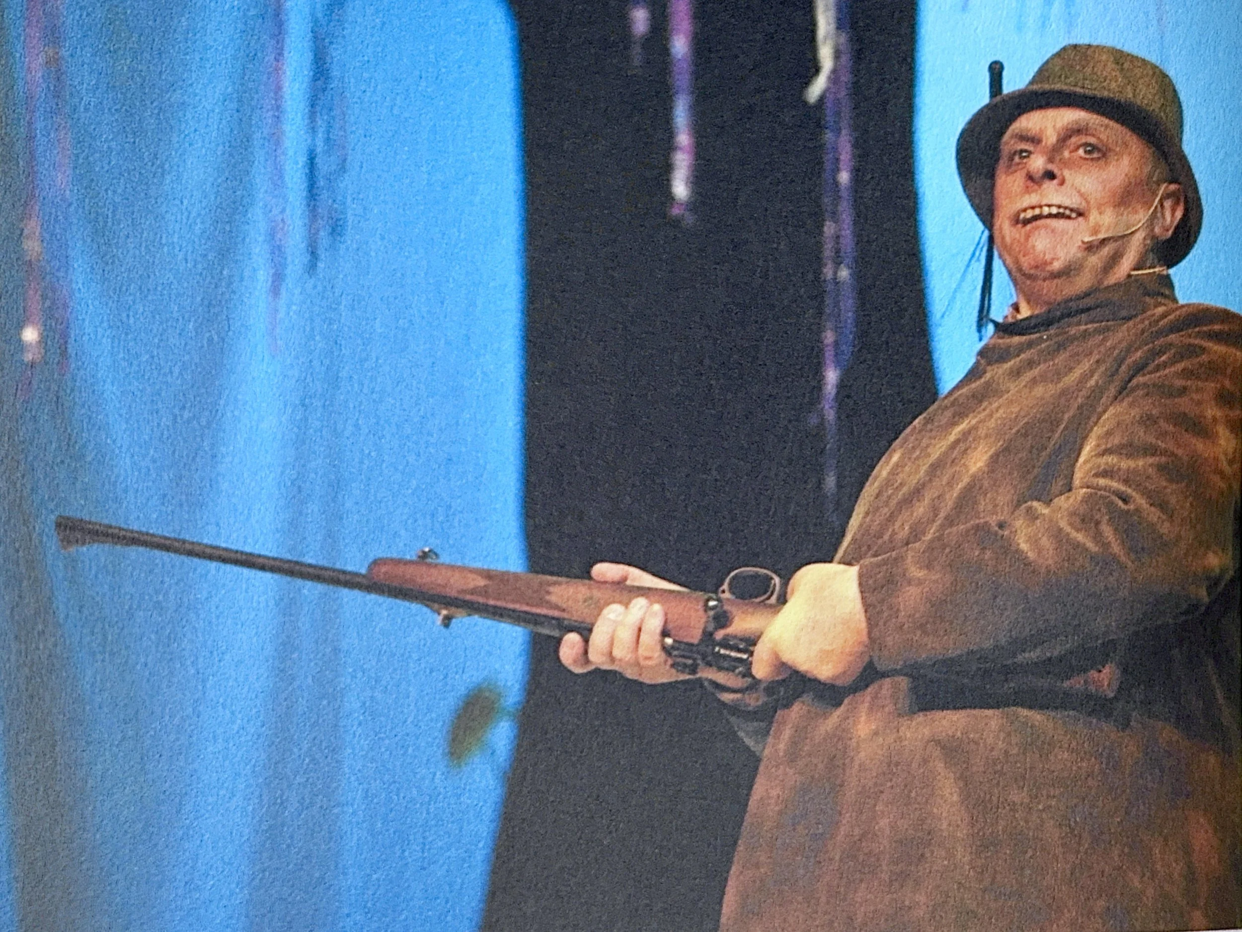 A man in a brown jacket and cowboy hat holds a rifle, standing on a stage with a blue background.