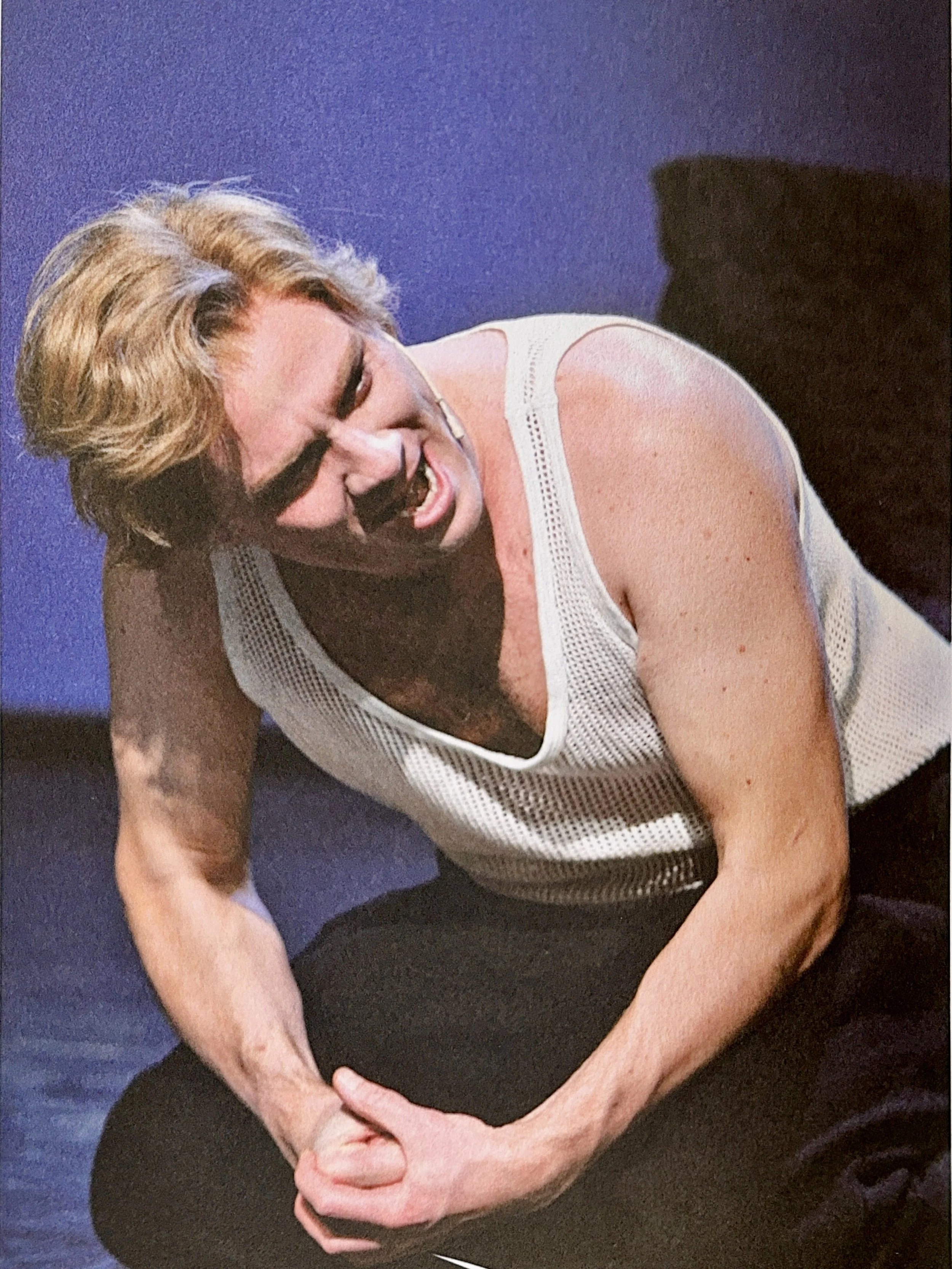 A person with blonde hair, wearing glasses and a white sleeveless top, sitting with one arm resting on their knee and their hand clasped over their stomach, against a dark blue background.