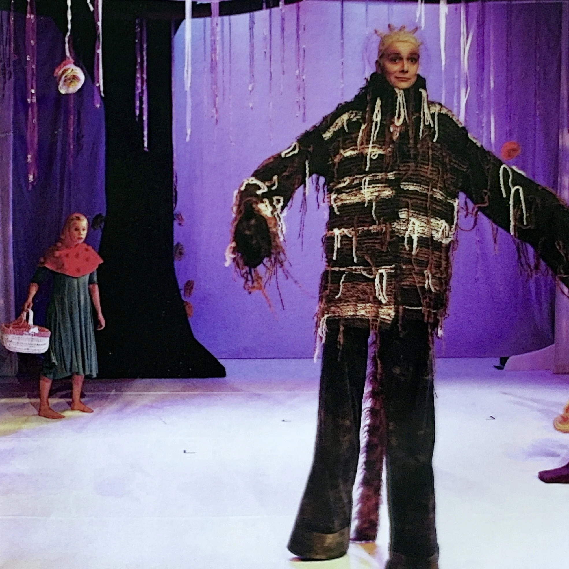 A person dressed in a colorful, striped costume with a hood, standing in a space with purple lighting. Two children are nearby, one in a red costume with a spotted pattern, and the other in a dark striped dress with yellow shoes, watching the perform