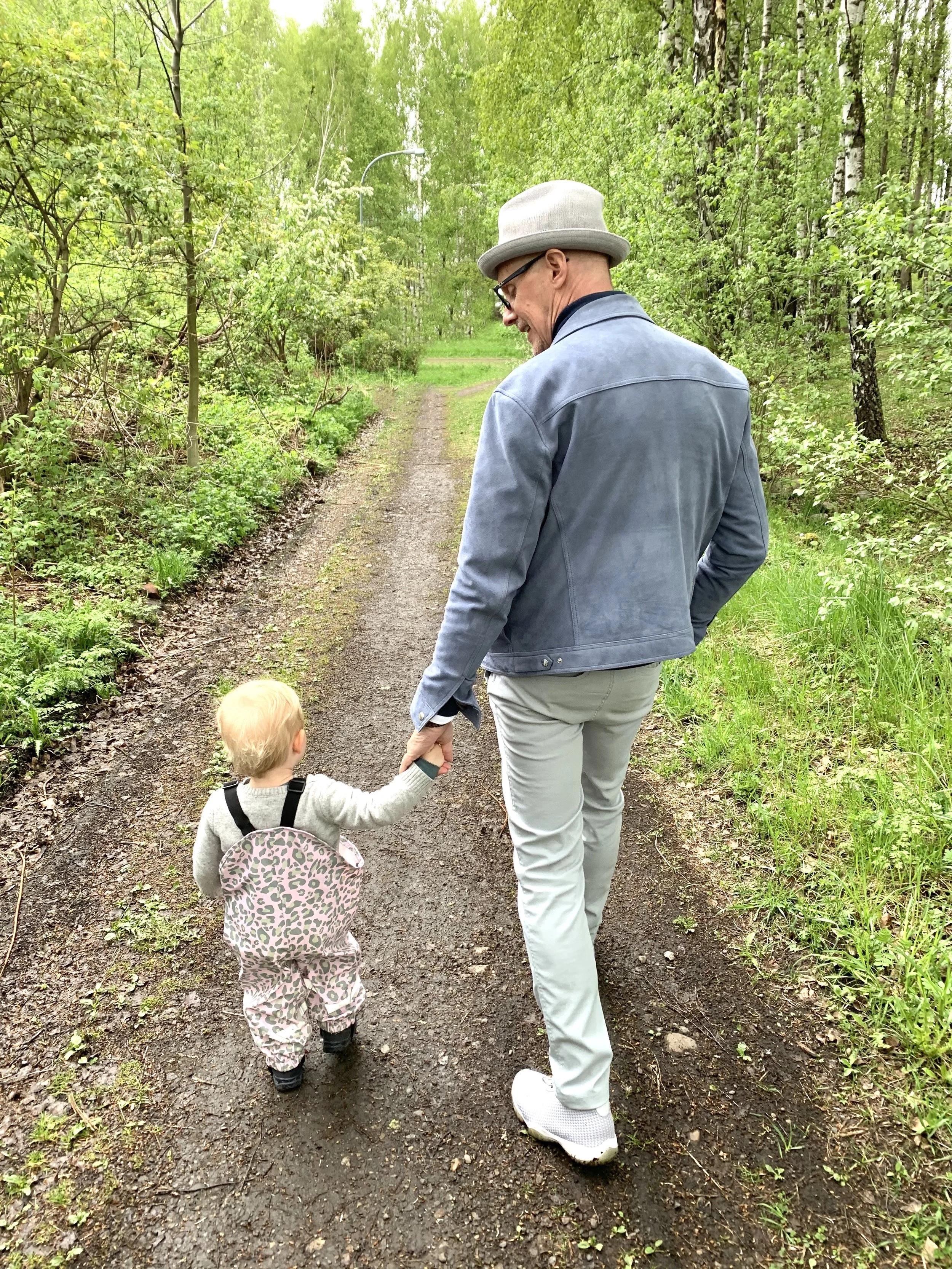 An adult man and a young girl hold hands while walking down a dirt trail in a lush green forest. The man is wearing a light gray hat, glasses, a blue jacket, and light-colored pants. The girl is dressed in patterned pants, a gray sweater, and black boots, and has a backpack with a pink and gray animal print.