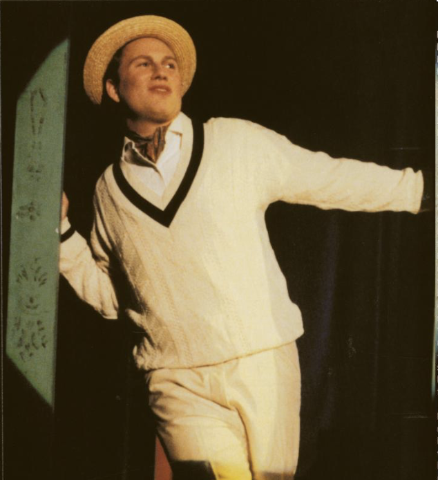A person dressed in vintage clothing, including a straw hat, sweater, and slacks, standing with one arm extended to the side.