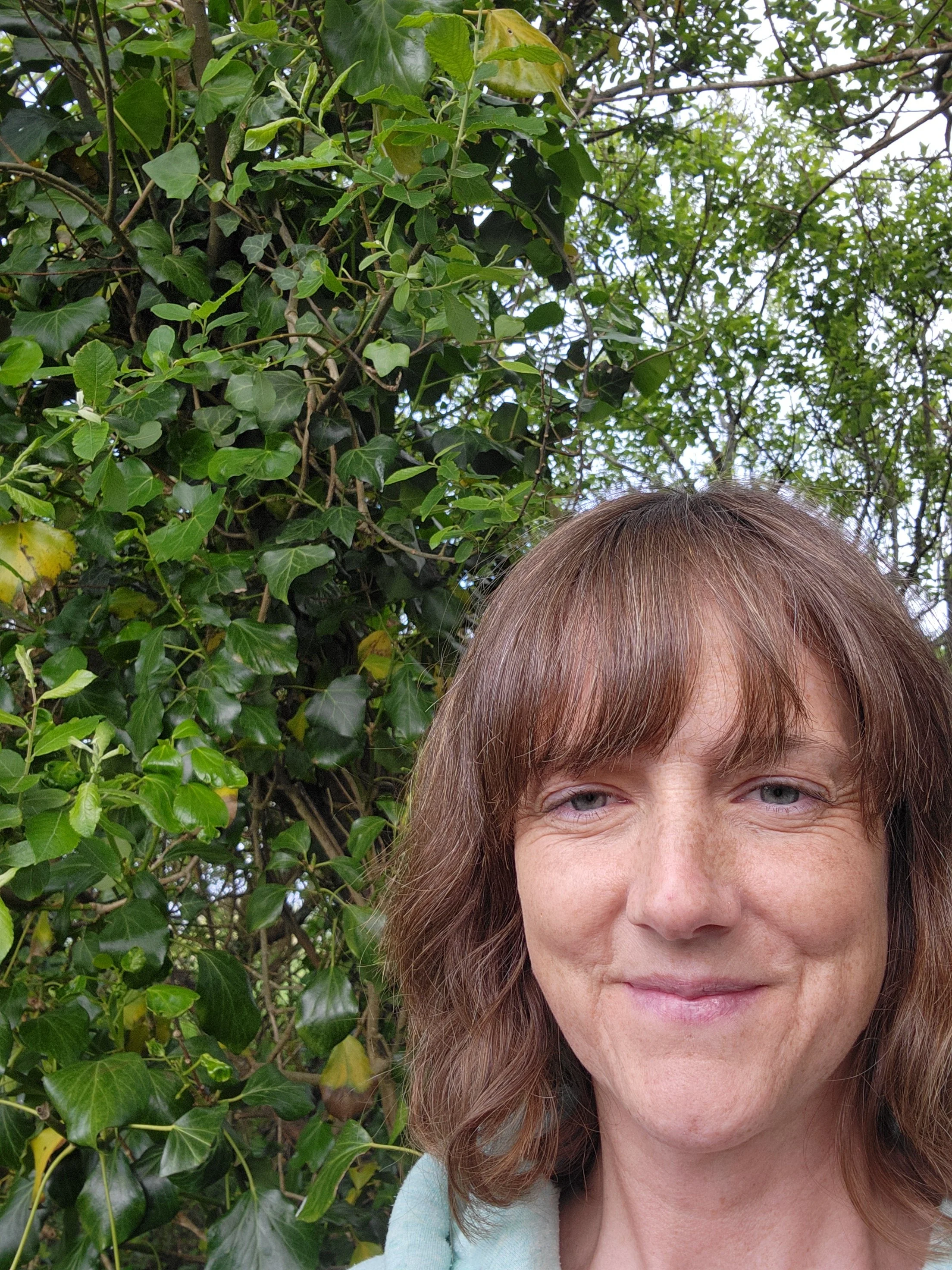 A woman with light skin and reddish-brown hair partially visible in a close-up photo, standing outdoors near green foliage, with a background of leafy trees.