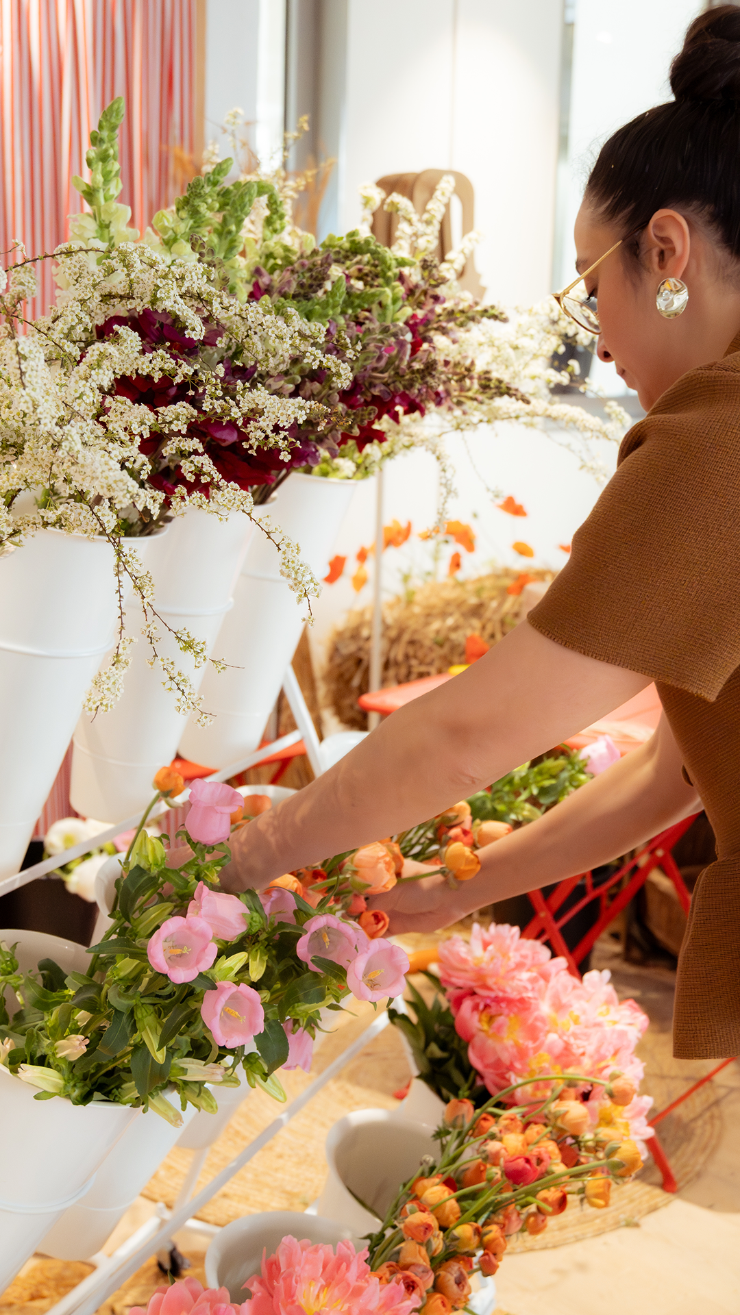 Une femme arrangeant des fleurs dans un magasin de fleurs, avec une étagère de fleurs colorées devant elle et un intérieur lumineux.