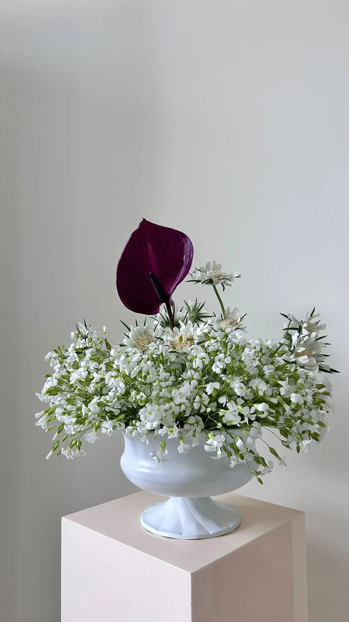 Un bouquet de fleurs blanches dans un vase blanc, avec une grande fleur violette en haut, placé sur un support blanc contre un fond gris clair.