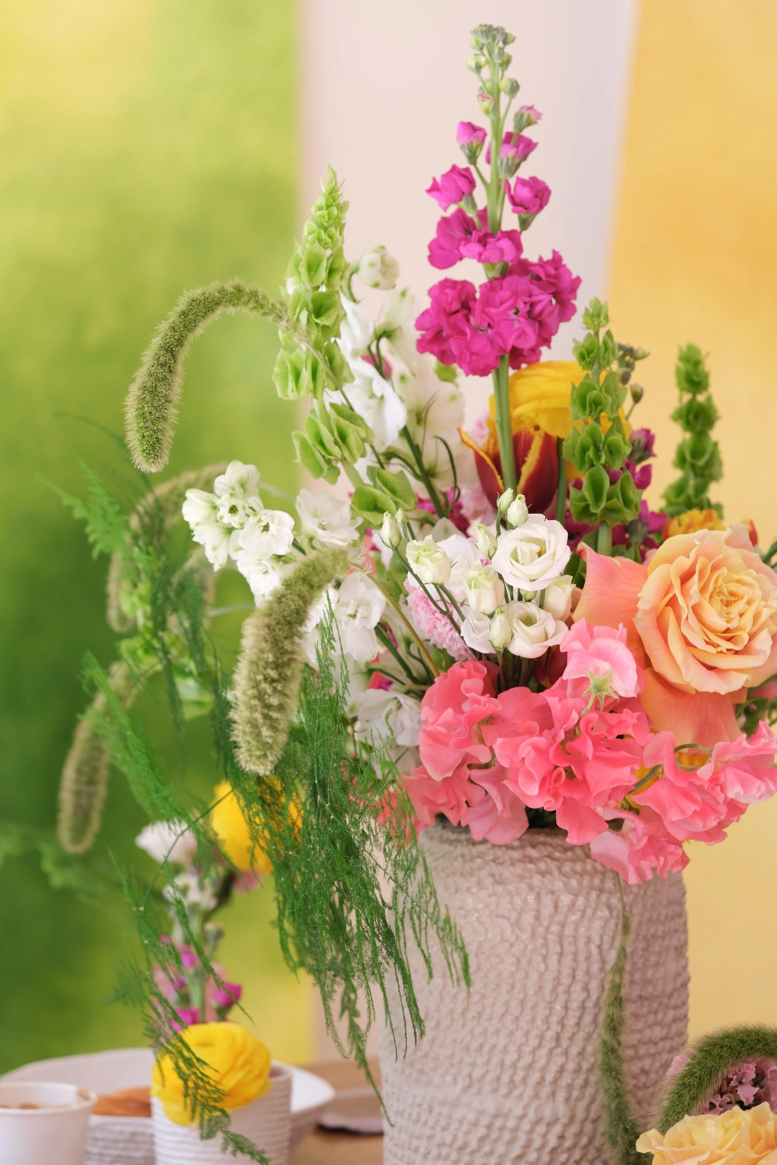 Un bouquet de fleurs colorées dans un vase en céramique texturé, avec des fleurs roses, blanches, jaunes et orange, placé sur une table dans une ambiance lumineuse.