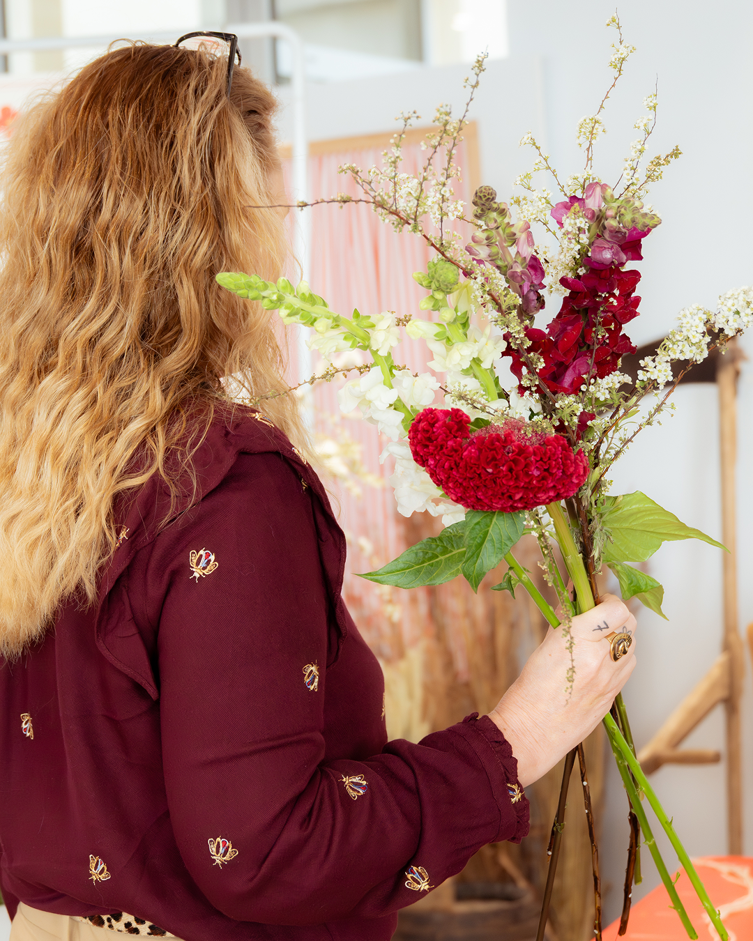 Femme tenant un bouquet de fleurs colorées, vue de côté, avec des cheveux rouges bouclés et lunettes sur la tête, dans un intérieur lumineux.