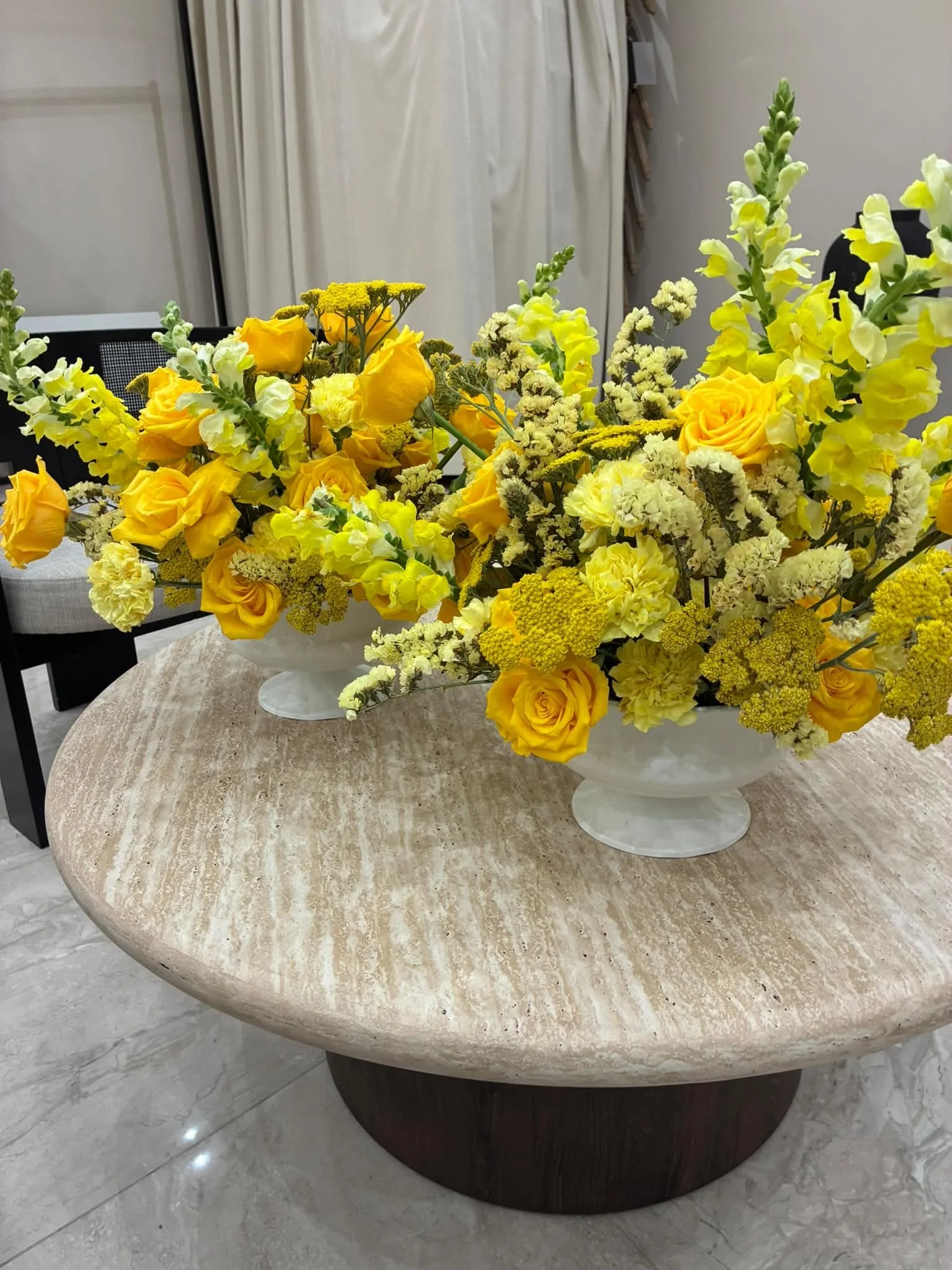 Un arrangement de fleurs jaunes et orange dans deux vases blancs placés sur une table en marbre beige, dans une pièce intérieure.