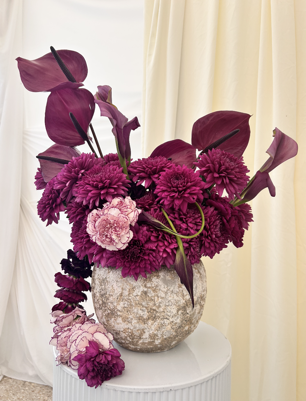 Un vase rempli de fleurs violettes, roses et mauves, avec des calas, des dahlias et des tournesols violets, placé sur une table blanche près de rideaux crème.