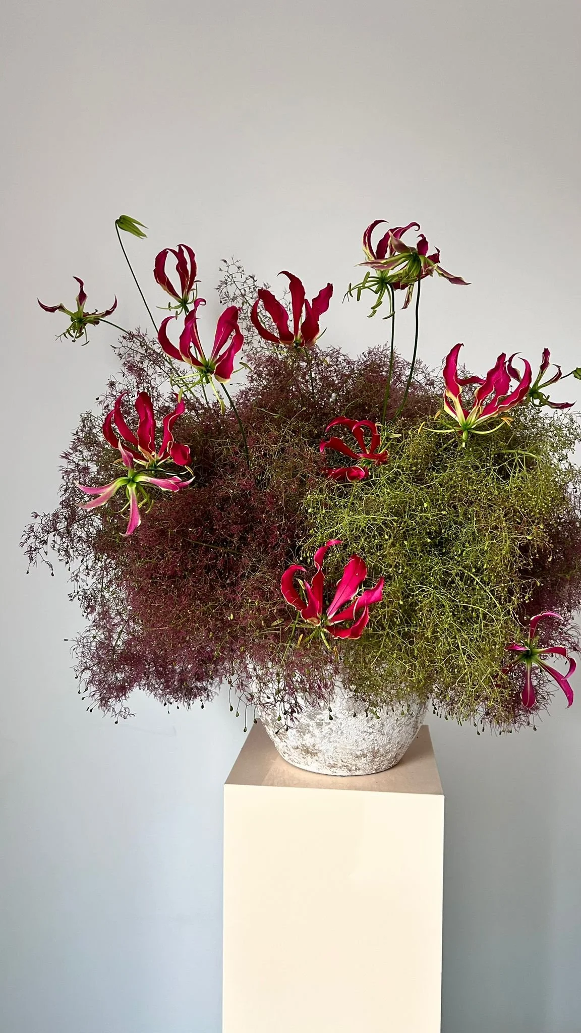 Un bouquet de fleurs roses et vertes dans un vase blanc sur une colonne blanche, contre un mur gris clair.