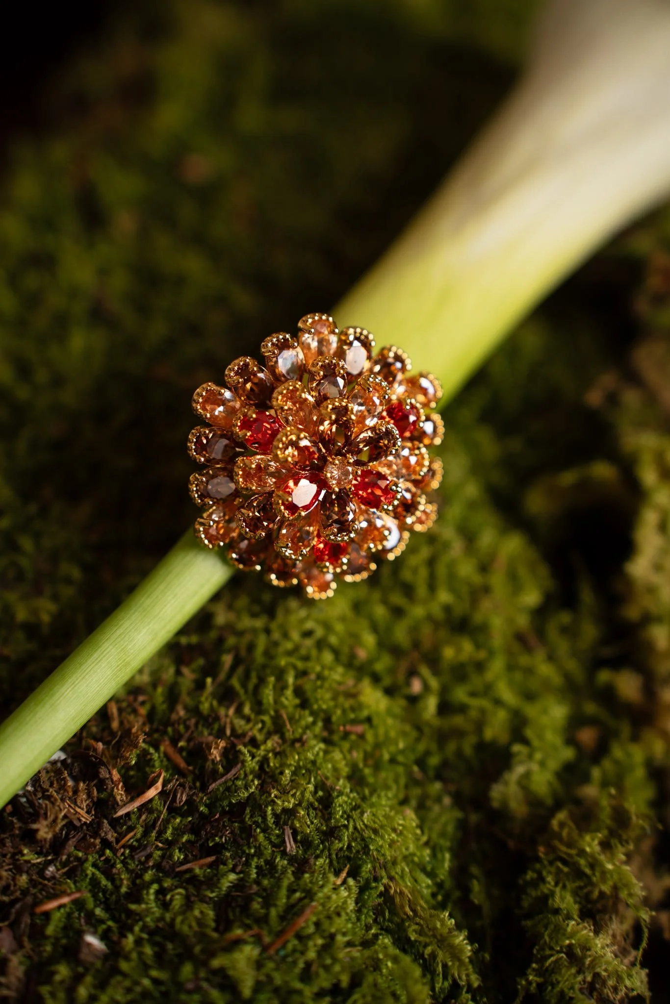 Un objet doré en forme de boule décorative ornée de pierres précieuses de couleur rouge et claire, posé sur un brin de plante verte avec un fond de mousse verte.
