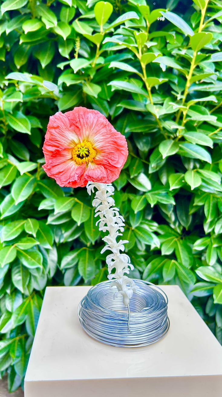 Une sculpture florale composée d'une grande fleur rouge avec un centre jaune, fixée à une structure blanche qui ressemble à une tige de fleurs, posée sur une surface blanche devant un buisson de feuilles vertes.