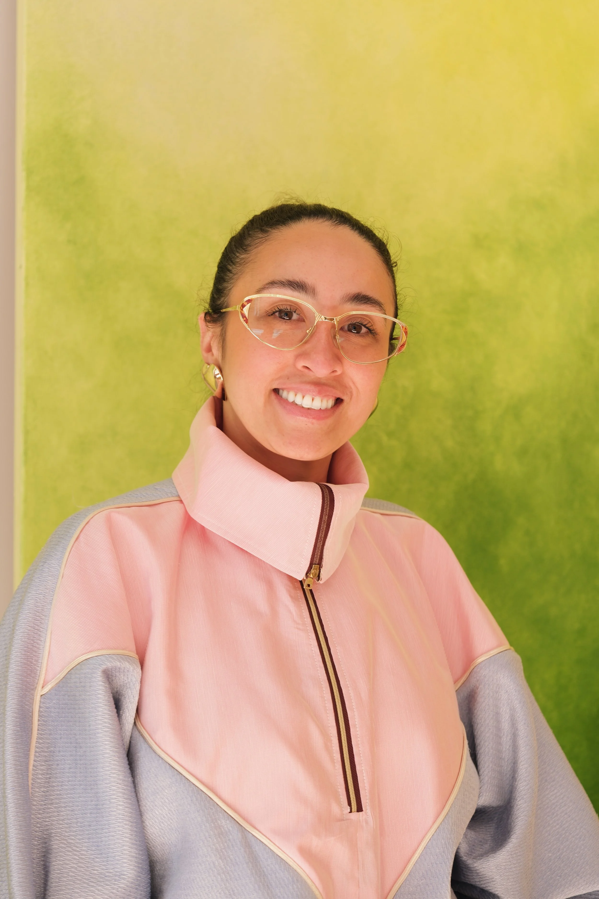 Jeune femme souriante portant des lunettes dorées, vêtue d'un sweat à mi-zip rose et gris clair, devant un mur vert. Elle a les cheveux attachés en arrière.