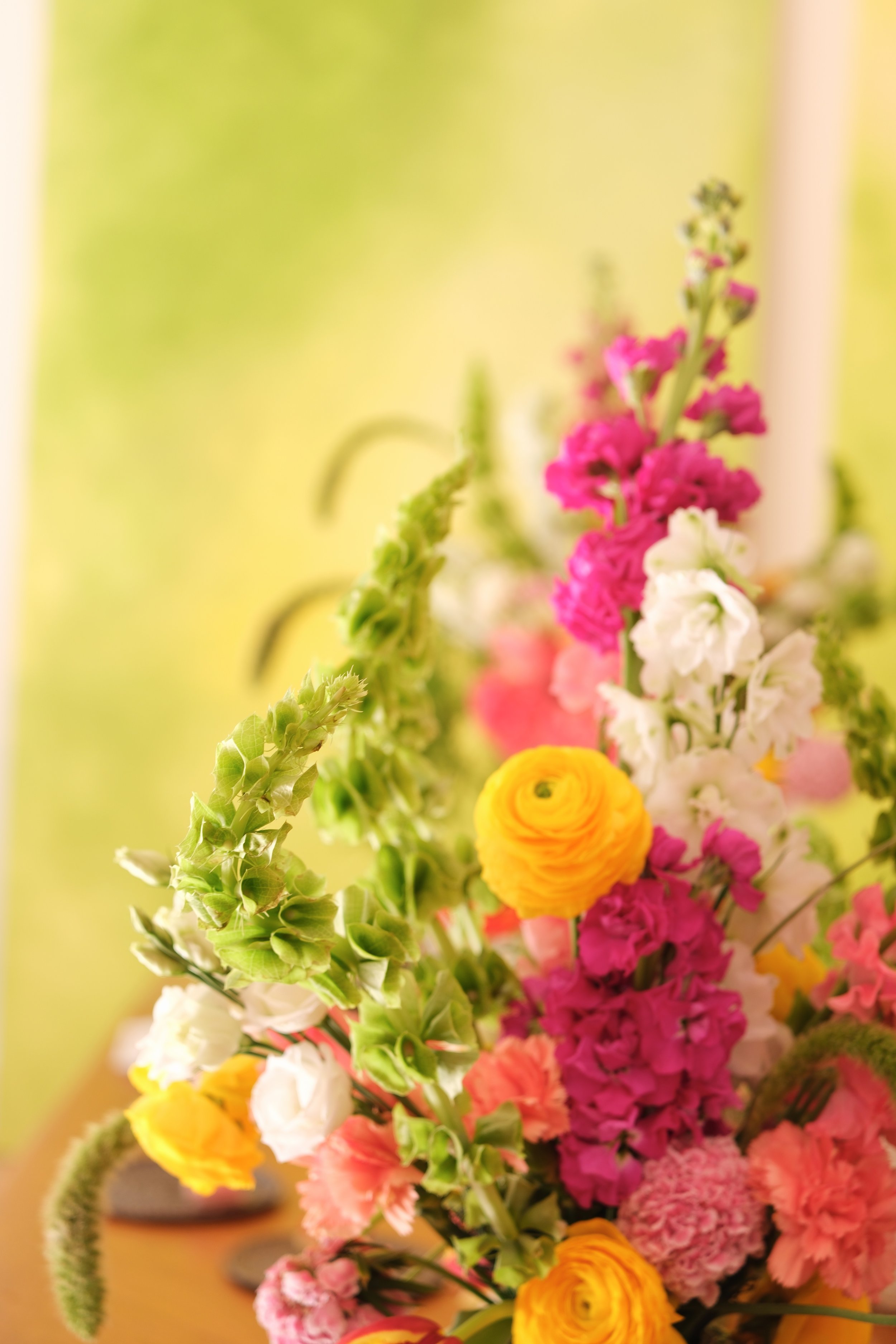 Un vase rempli de fleurs colorées, notamment des roses, des œillets et des statices, placé sur une table en bois, avec un fond flou vert.