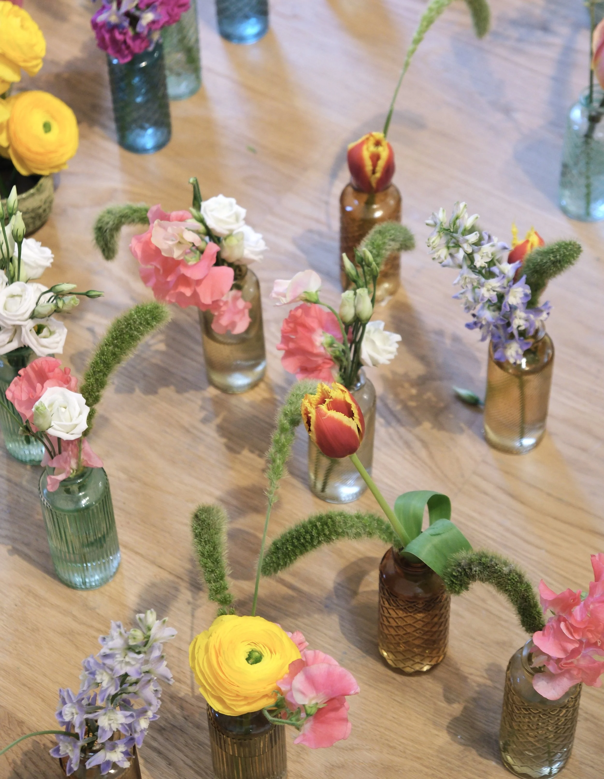 Une table en bois avec plusieurs vases en verre contenant des fleurs colorées, y compris des tulipes, des roses, des freesias, et d'autres fleurs de différentes couleurs.